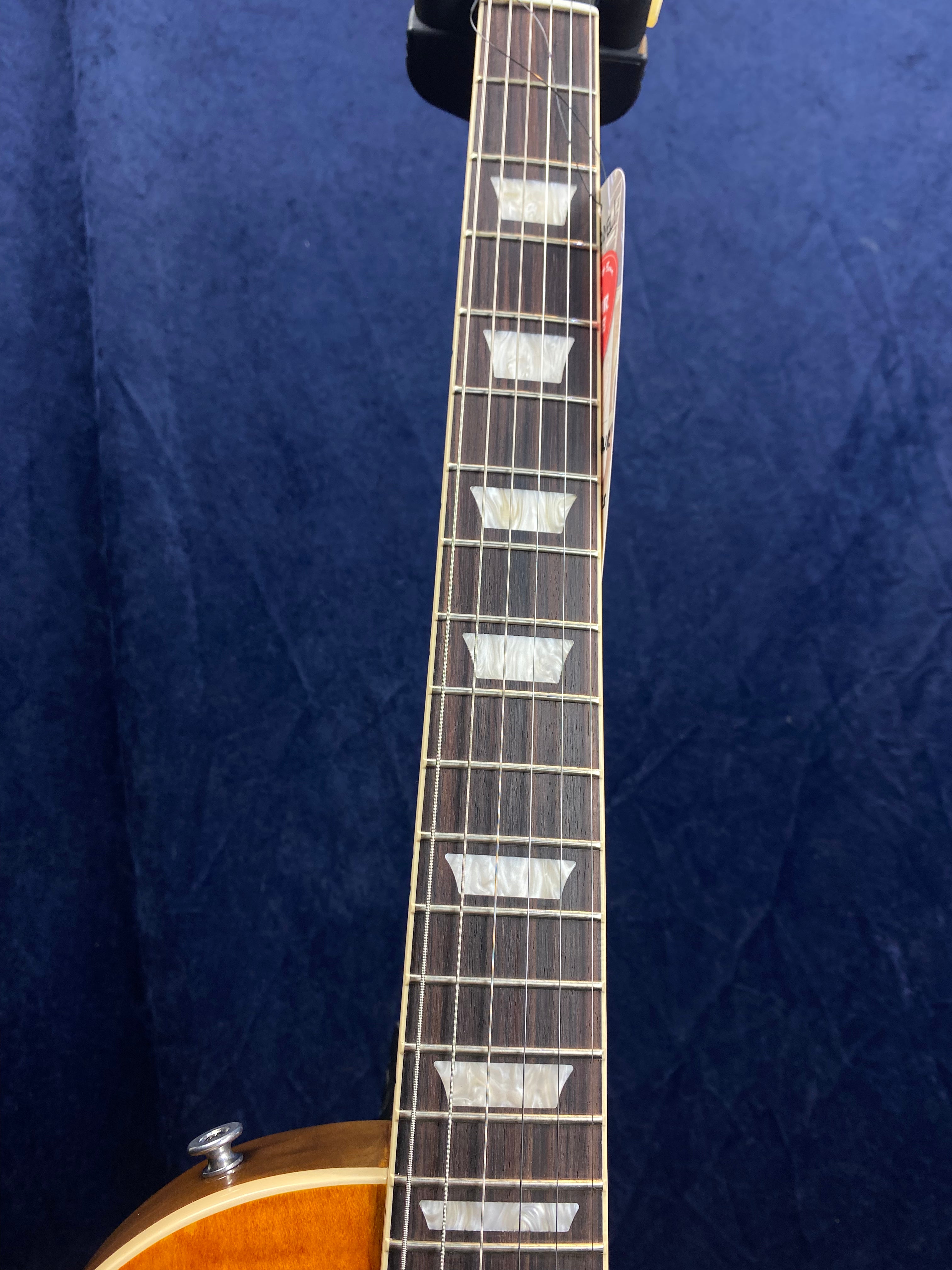 A close-up of an electric guitar neck with trapezoid inlays on a dark fingerboard, set against a dark blue background.