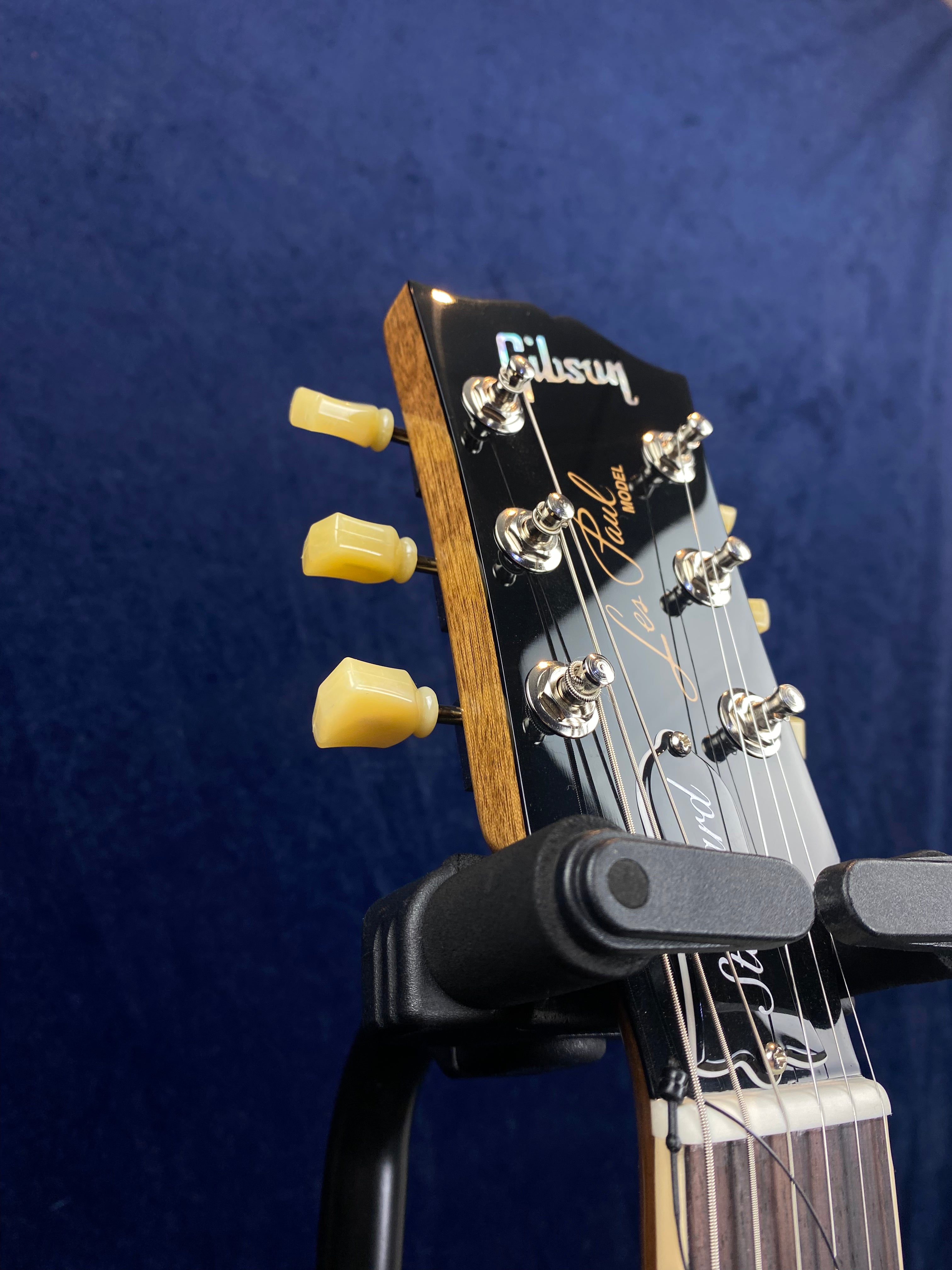 Headstock of a Gibson electric guitar with cream tuning pegs, metal tuning posts, and visible signature script, resting on a black guitar stand.