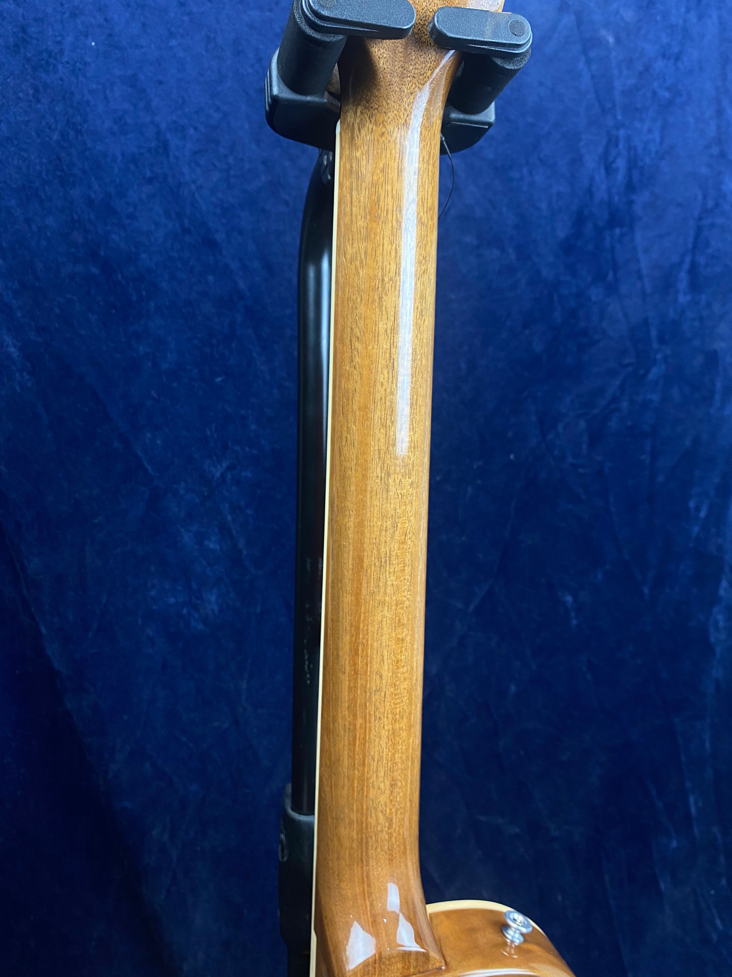 Glossy wooden electric guitar neck and headstock held upright in a black stand, blue textured background.
