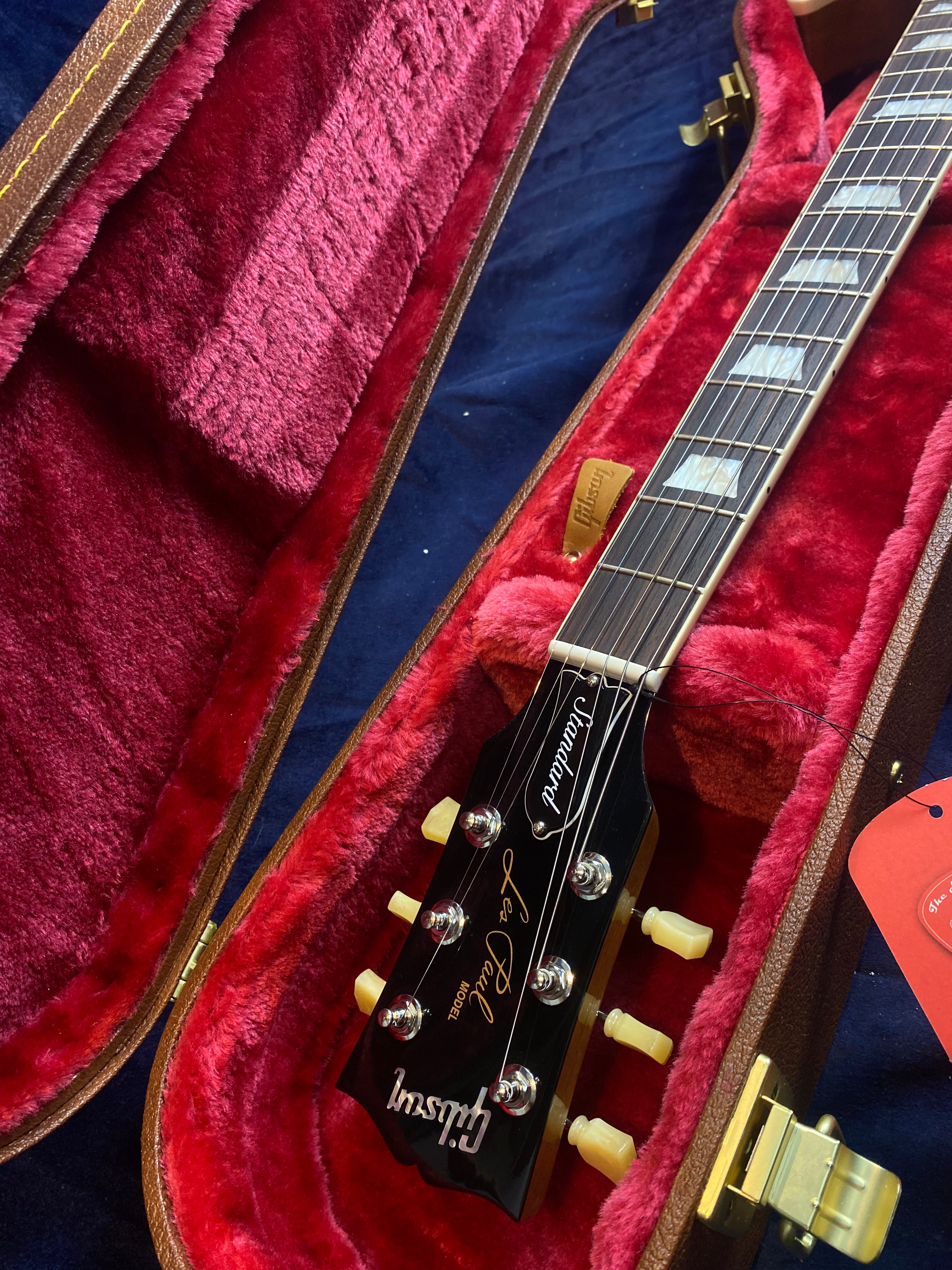 Gibson Les Paul electric guitar in a brown hard case lined with plush red fabric, showing the headstock, tuners, and part of the neck.