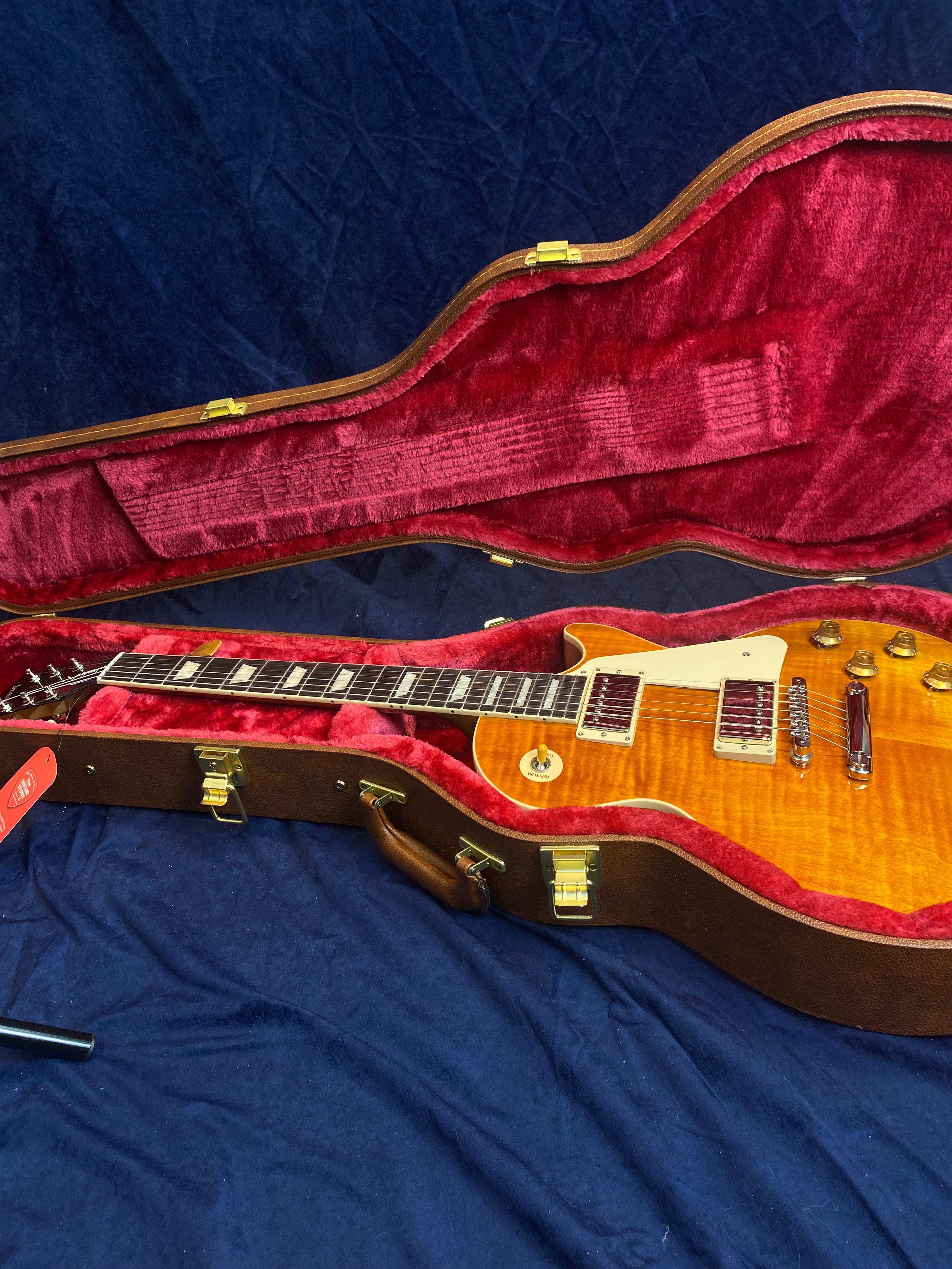 Sunburst electric guitar with cream pickguard rests in a plush red-lined hard case with gold latches, placed on a dark blue fabric surface.
