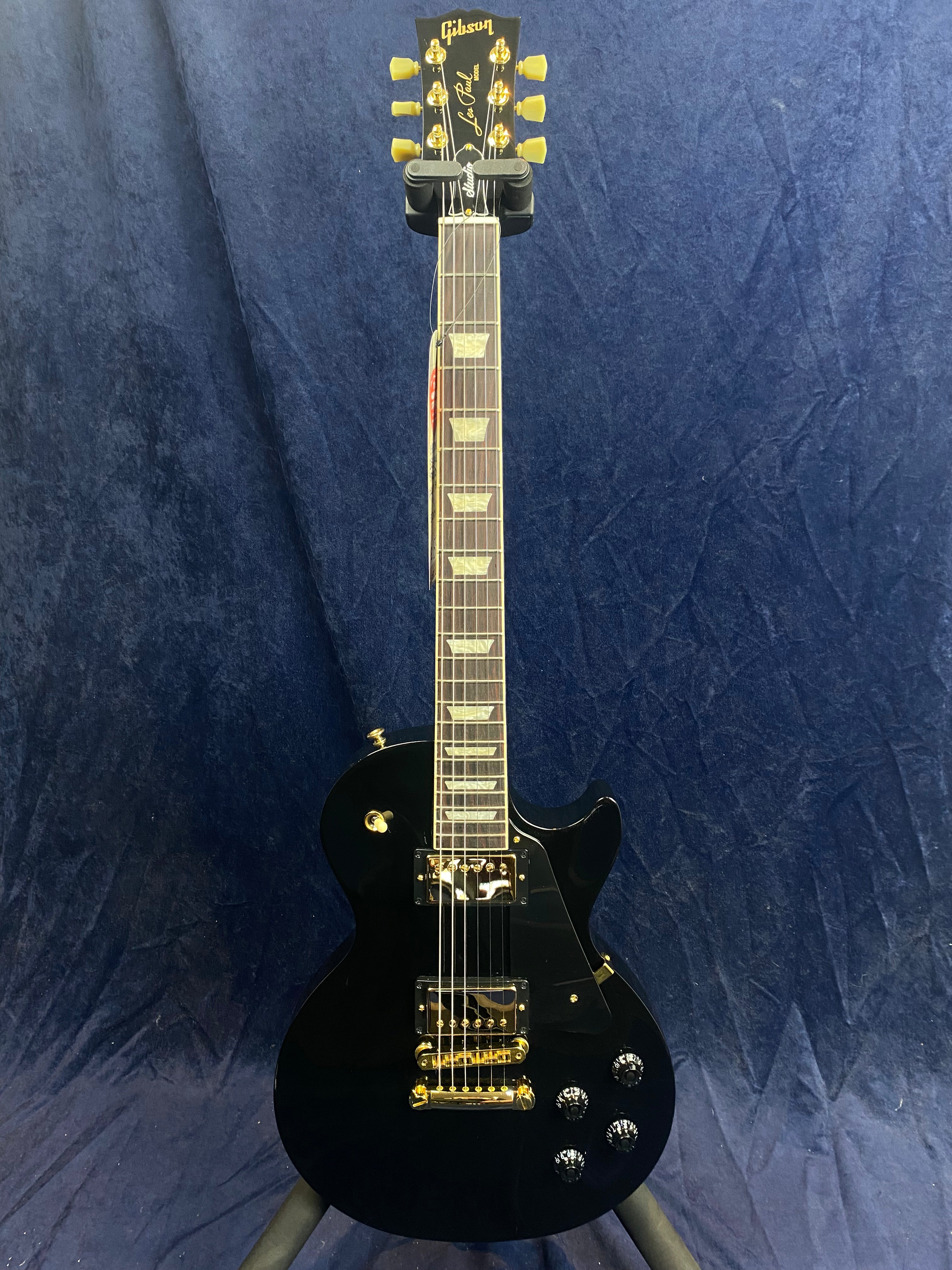 Black Gibson Les Paul electric guitar with gold hardware and trapezoid inlays, standing upright on a stand against a dark blue background.