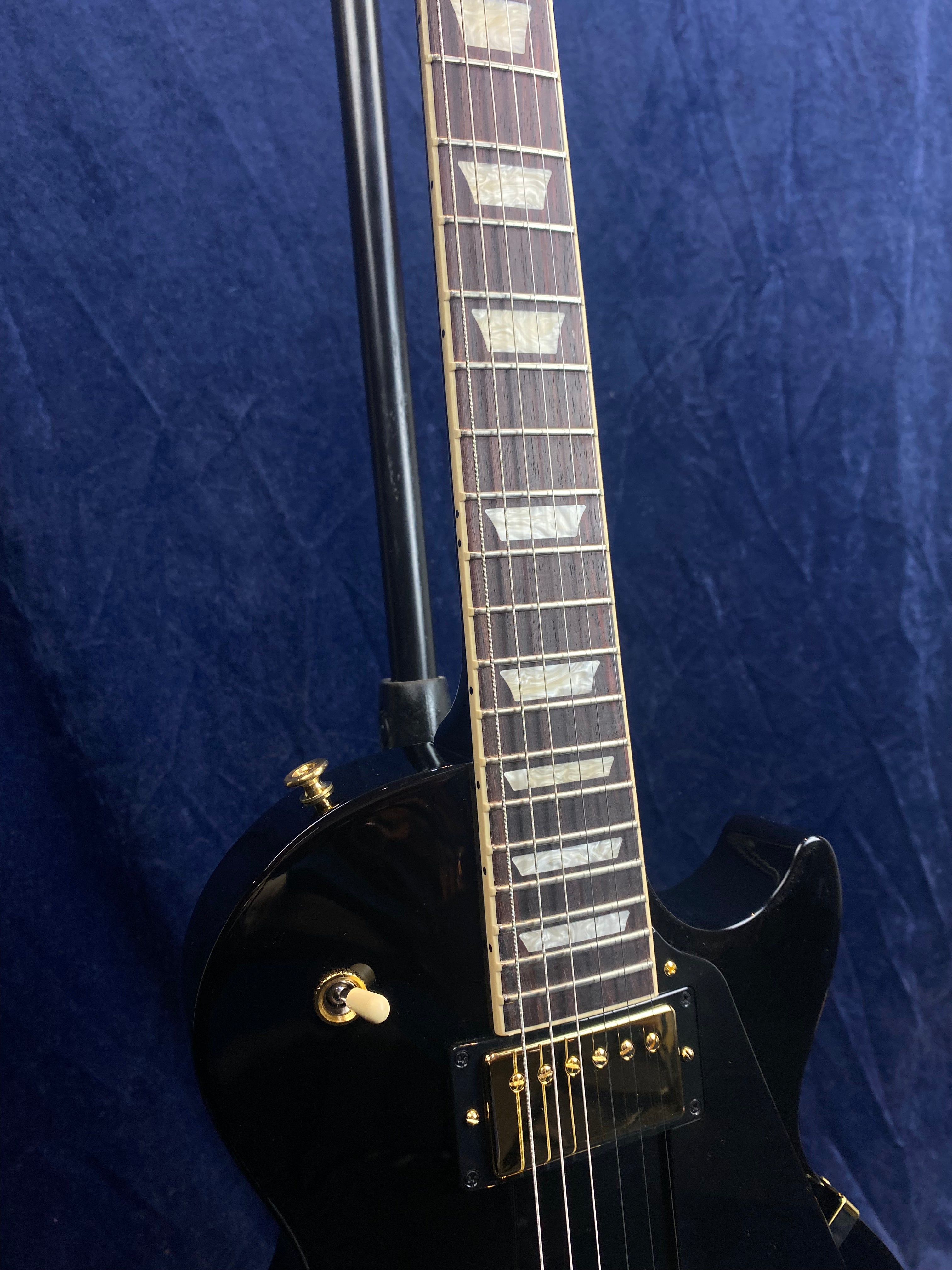 Black electric guitar with gold hardware and cream trapezoid inlays on a rosewood fretboard, shown partially against a dark blue background.
