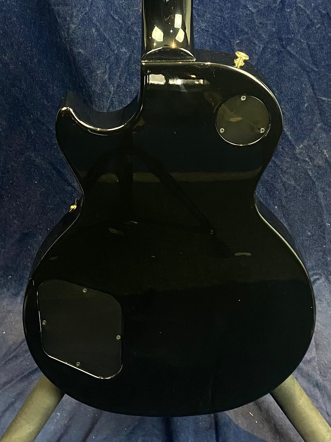 Glossy black electric guitar body, rear view, with two access panels and a gold strap button against a dark blue fabric background.
