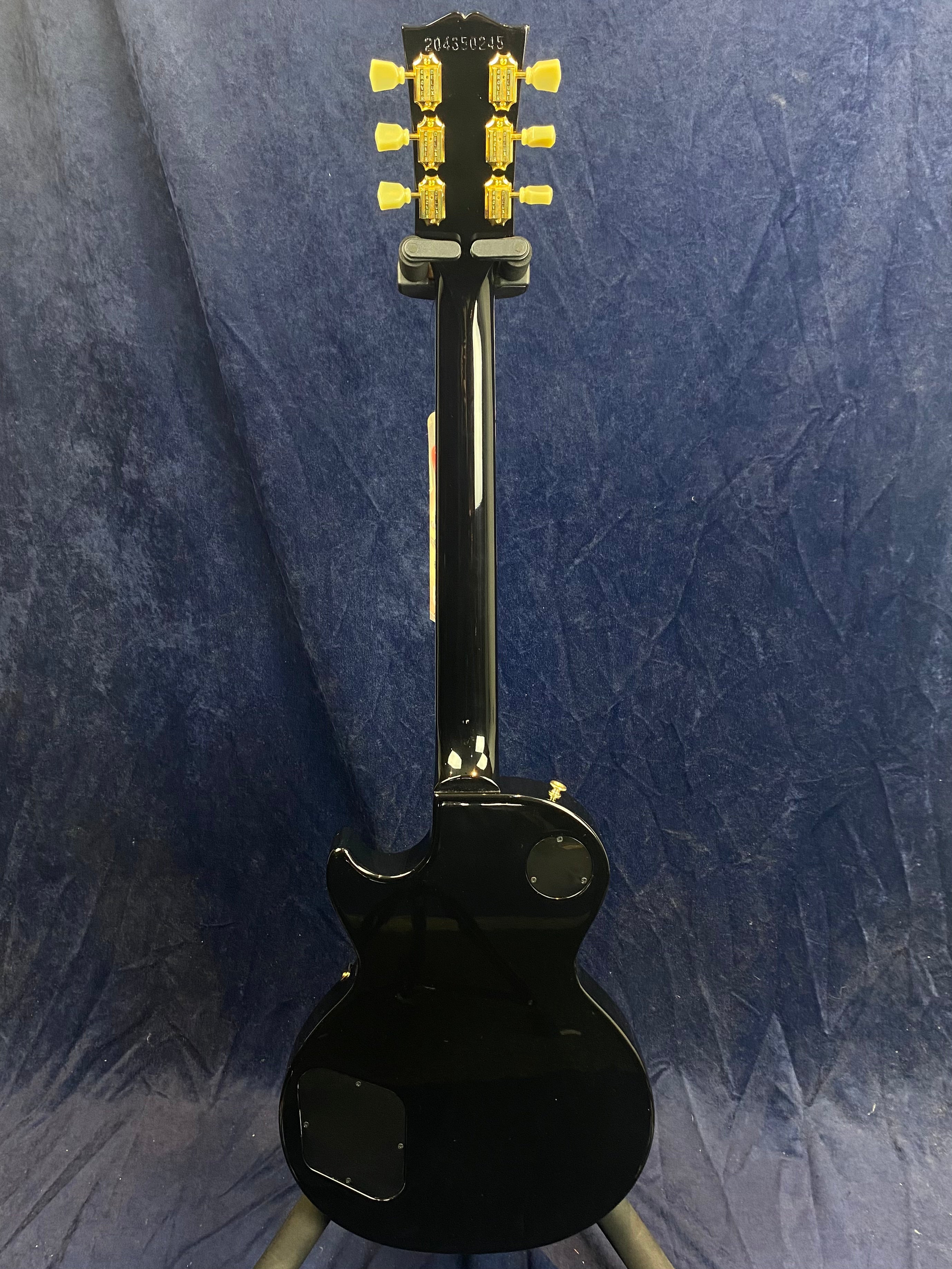 Black electric guitar seen from the back, gold tuning pegs, rounded single-cut body, control cavity covers visible, against a dark blue cloth background.