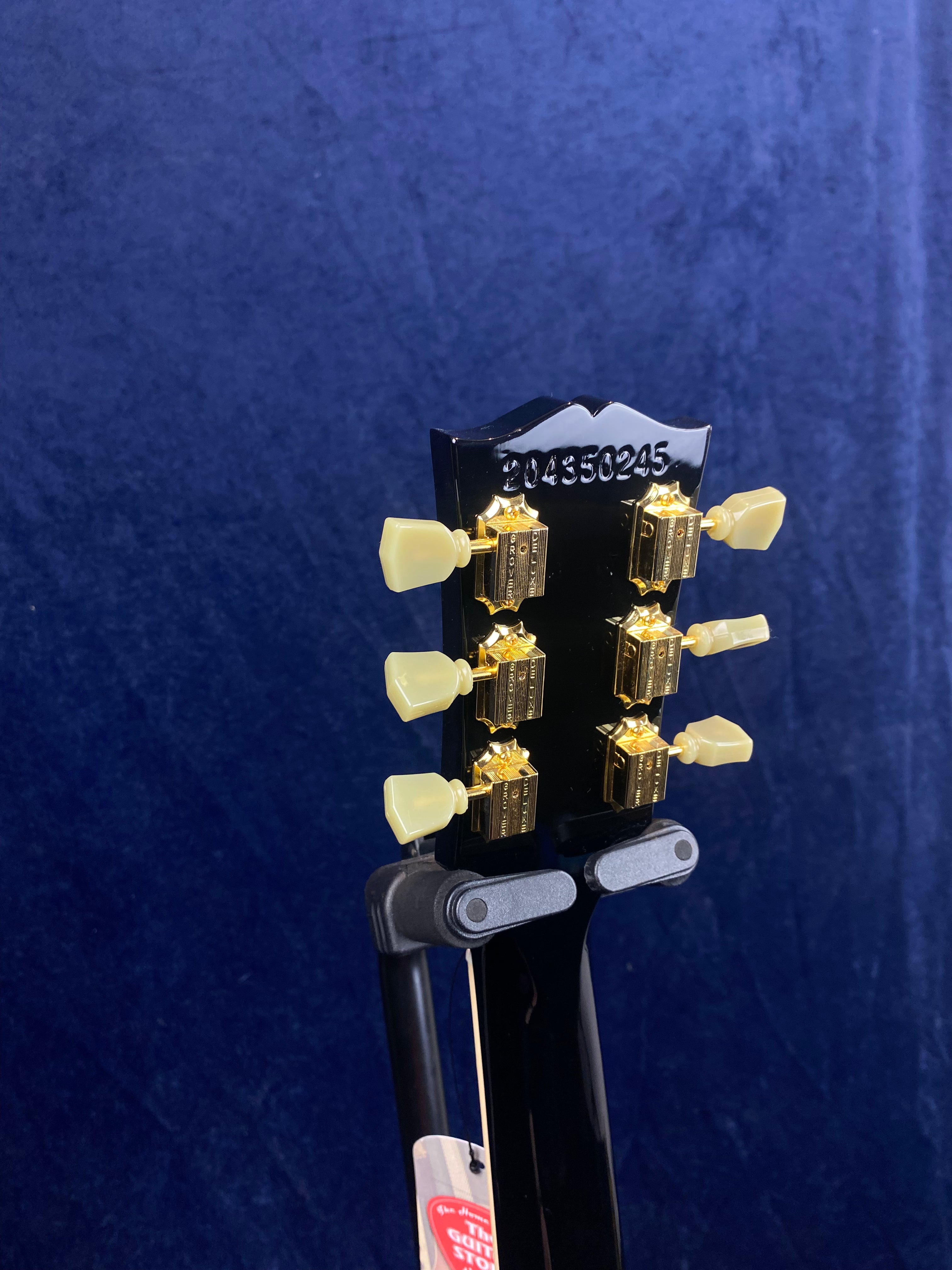 Back of an electric guitar headstock with gold tuning pegs and cream knobs, serial number 204350245 stamped at the top, against a blue background.