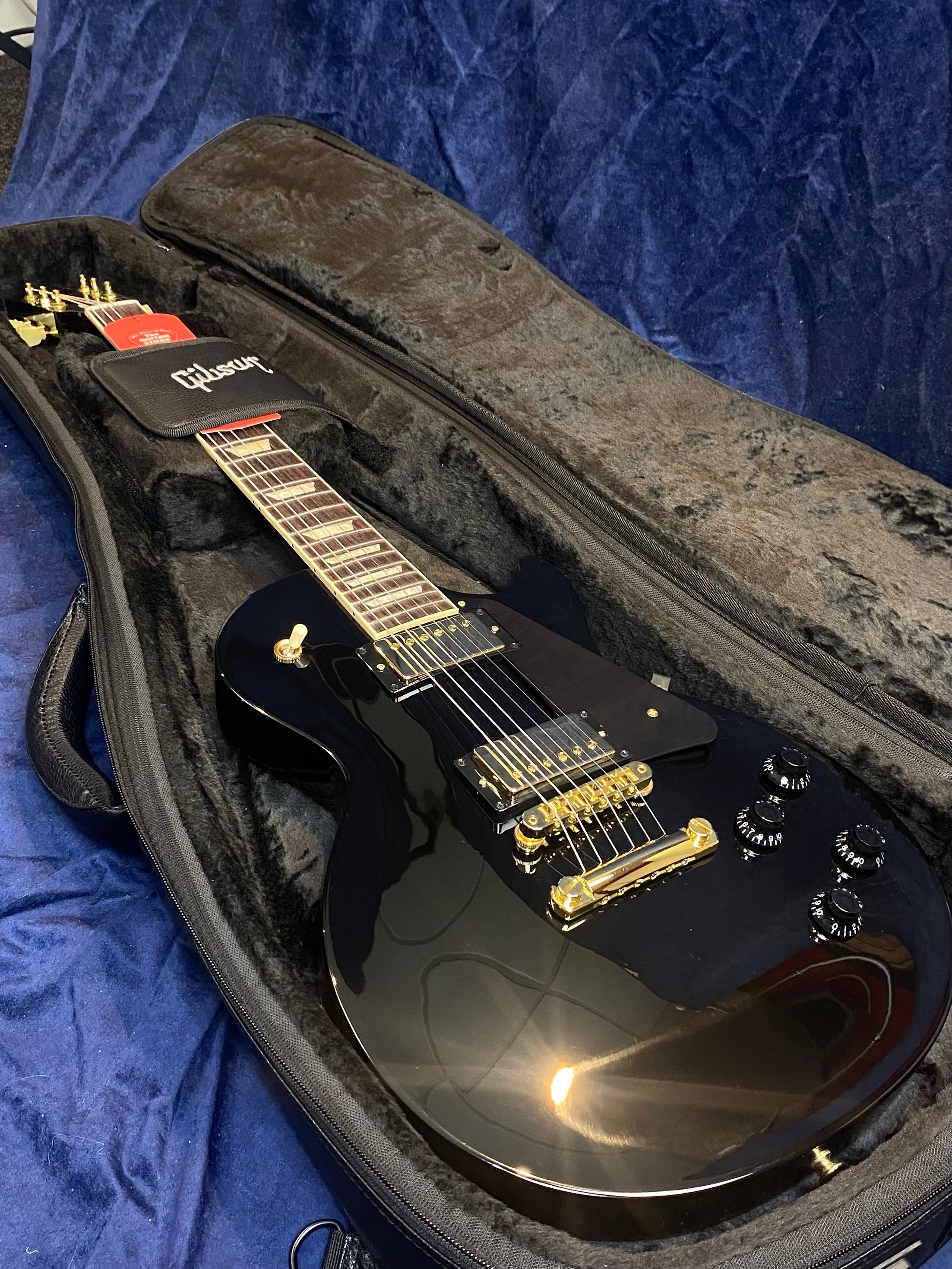 Black electric guitar with gold hardware and four black control knobs, resting inside a padded Gibson-branded case on a blue surface.
