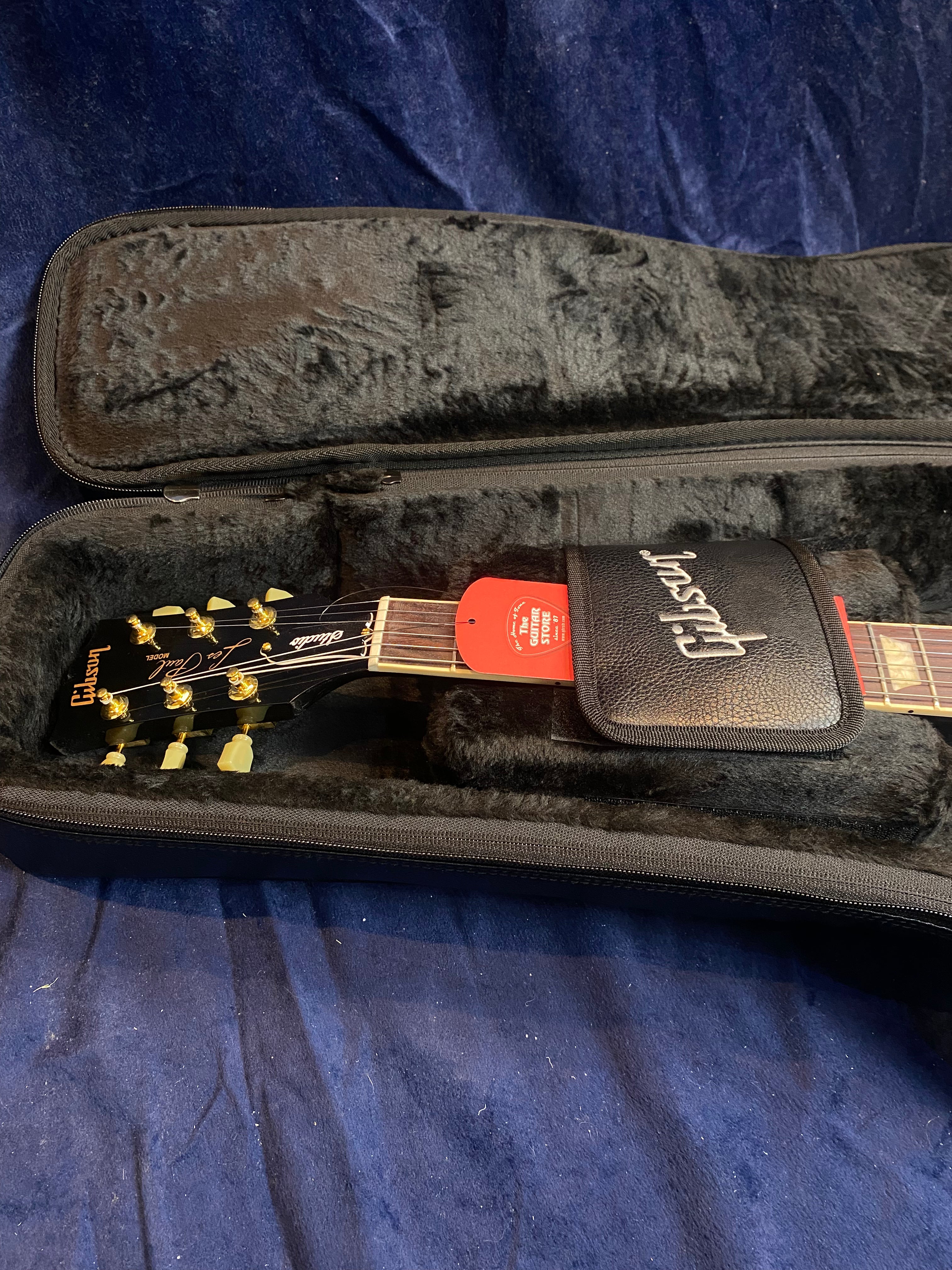 A Gibson electric guitar rests in a plush black case, showing the headstock with gold tuning pegs and a red tag attached to the neck.