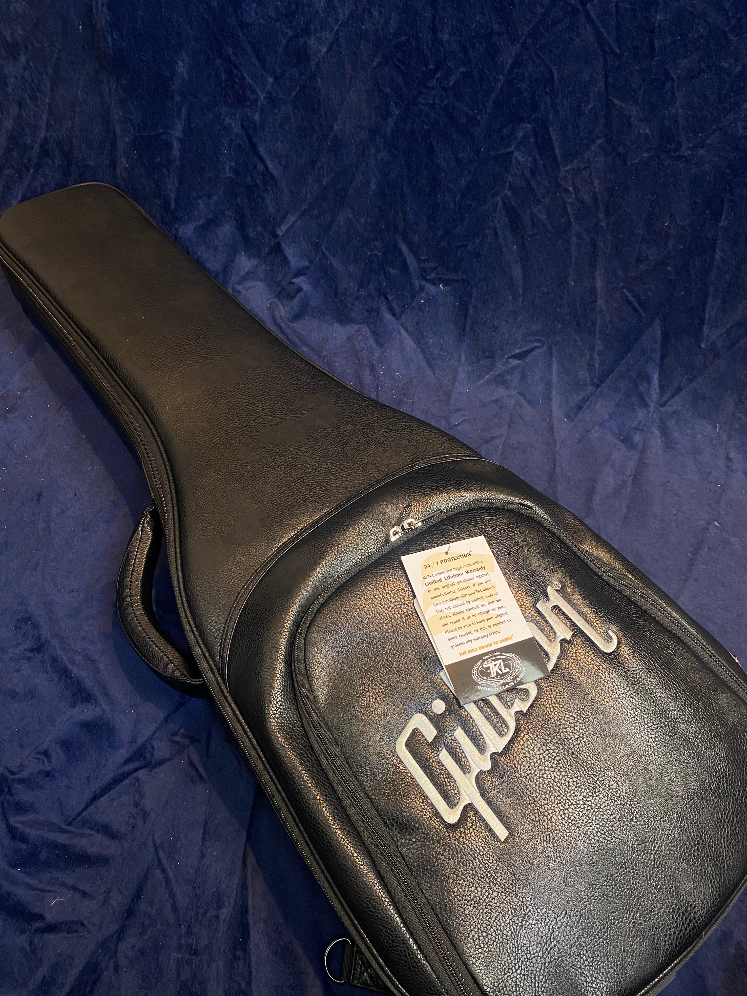 Black Gibson-branded electric guitar gig bag on a dark blue textured background, with a warranty tag attached to the front pocket zipper.