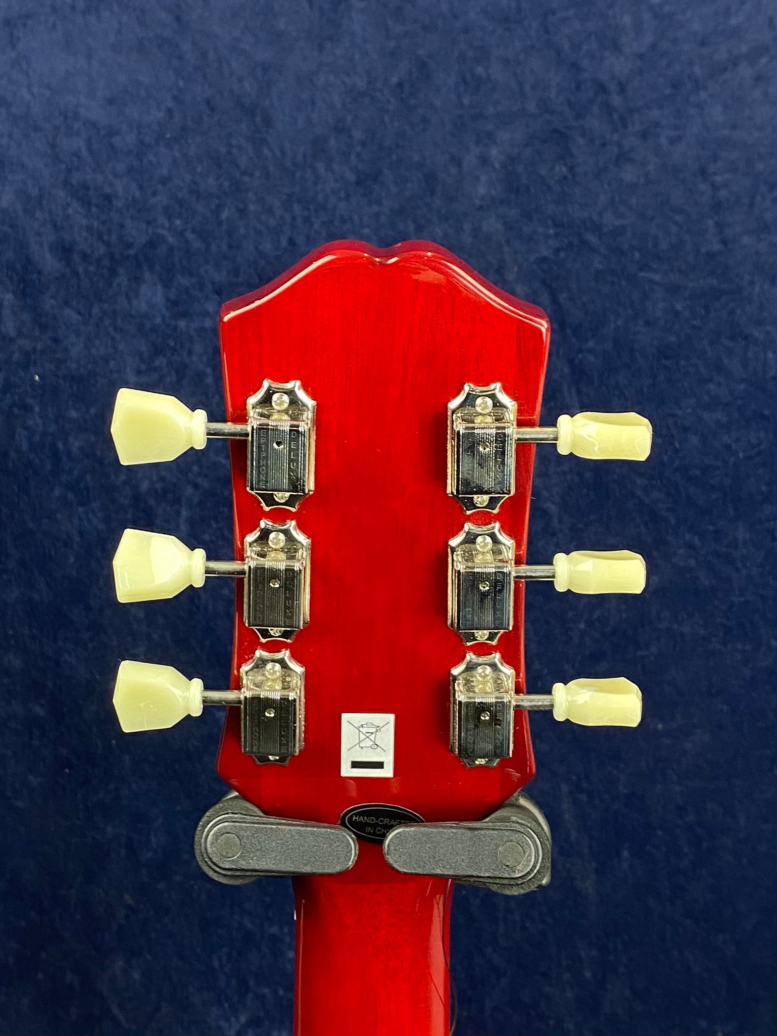 Red electric guitar headstock with six cream tuning pegs and metal tuners, "hand-crafted in China" label at the base, blue background.