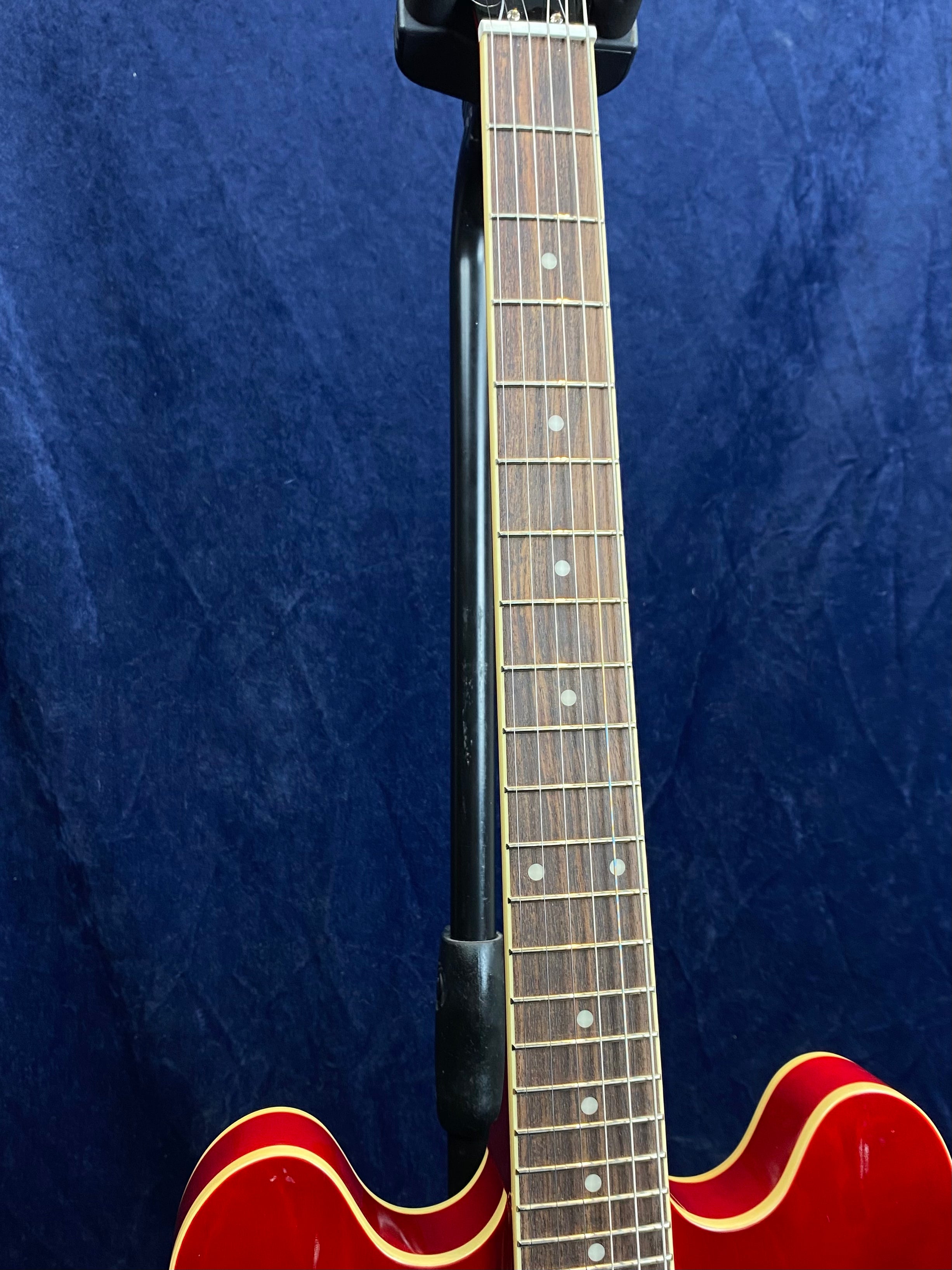 Red electric guitar with a wooden fretboard, white dot inlays, and cream binding, hanging on a black wall-mounted holder against a dark blue background.