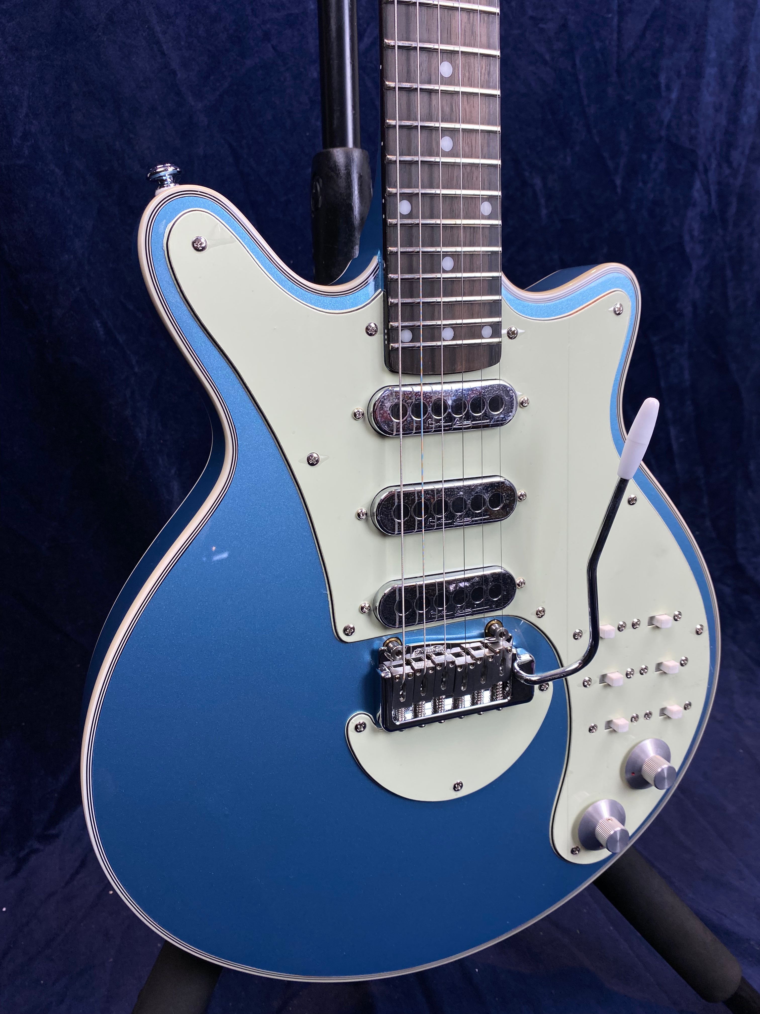 Metallic blue electric guitar with a white pickguard, three pickups, white control knobs, and a tremolo arm, shown on a stand against a dark background.