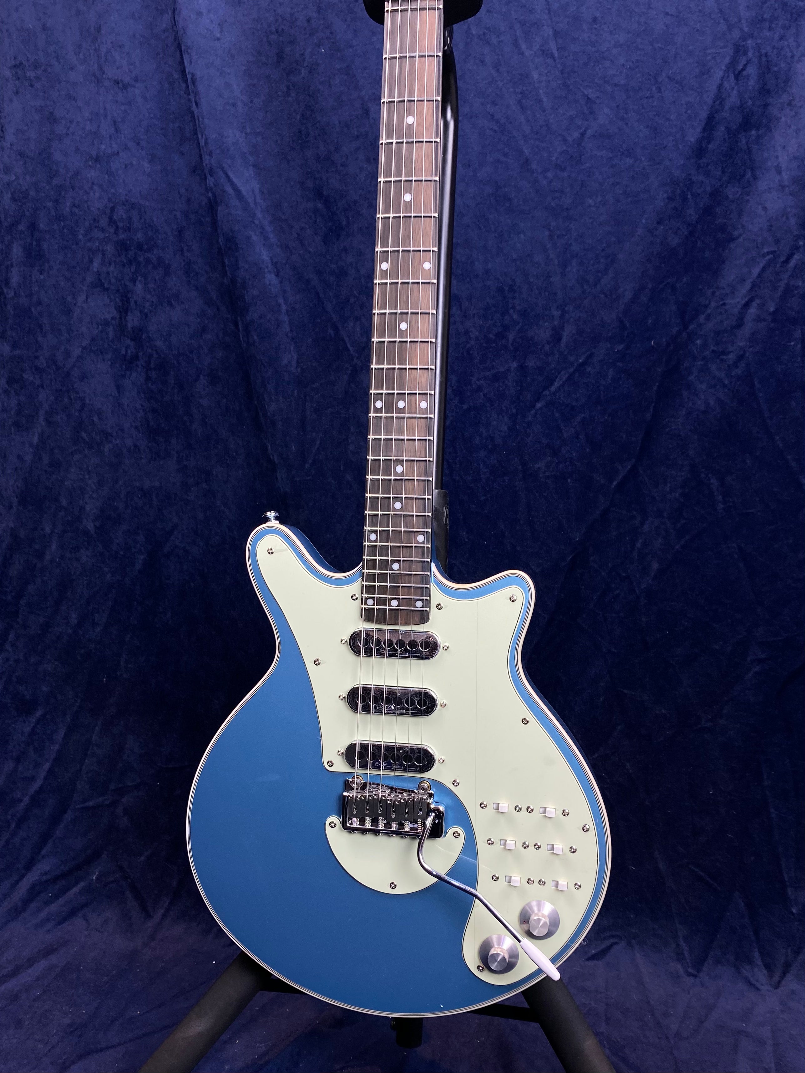 Blue electric guitar with three black pickups, white pickguard, multiple switches, two silver knobs, and a white tremolo bar on a stand.