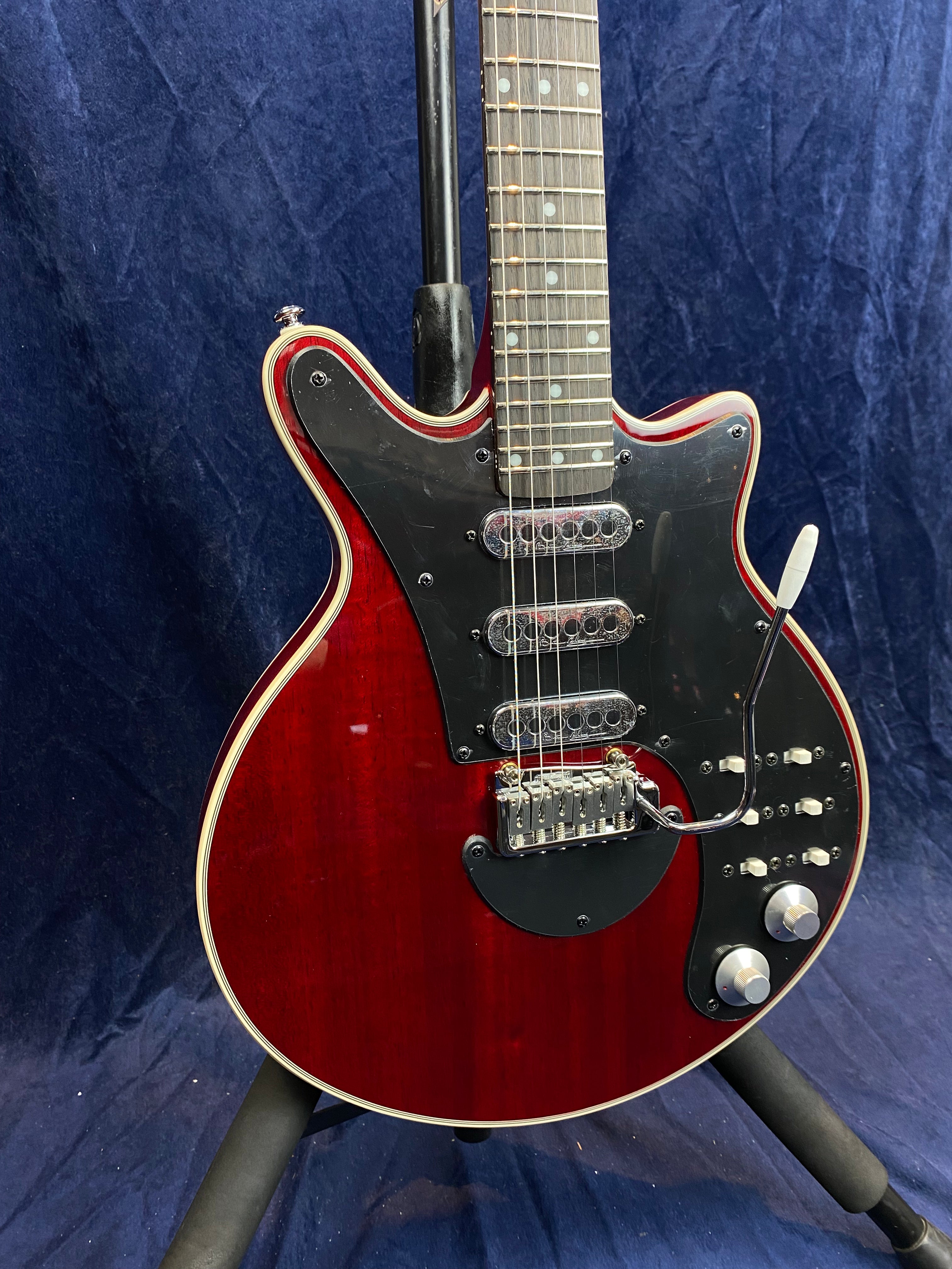 Red electric guitar with a black pickguard, three single-coil pickups, multiple switches, and silver control knobs, resting on a stand.