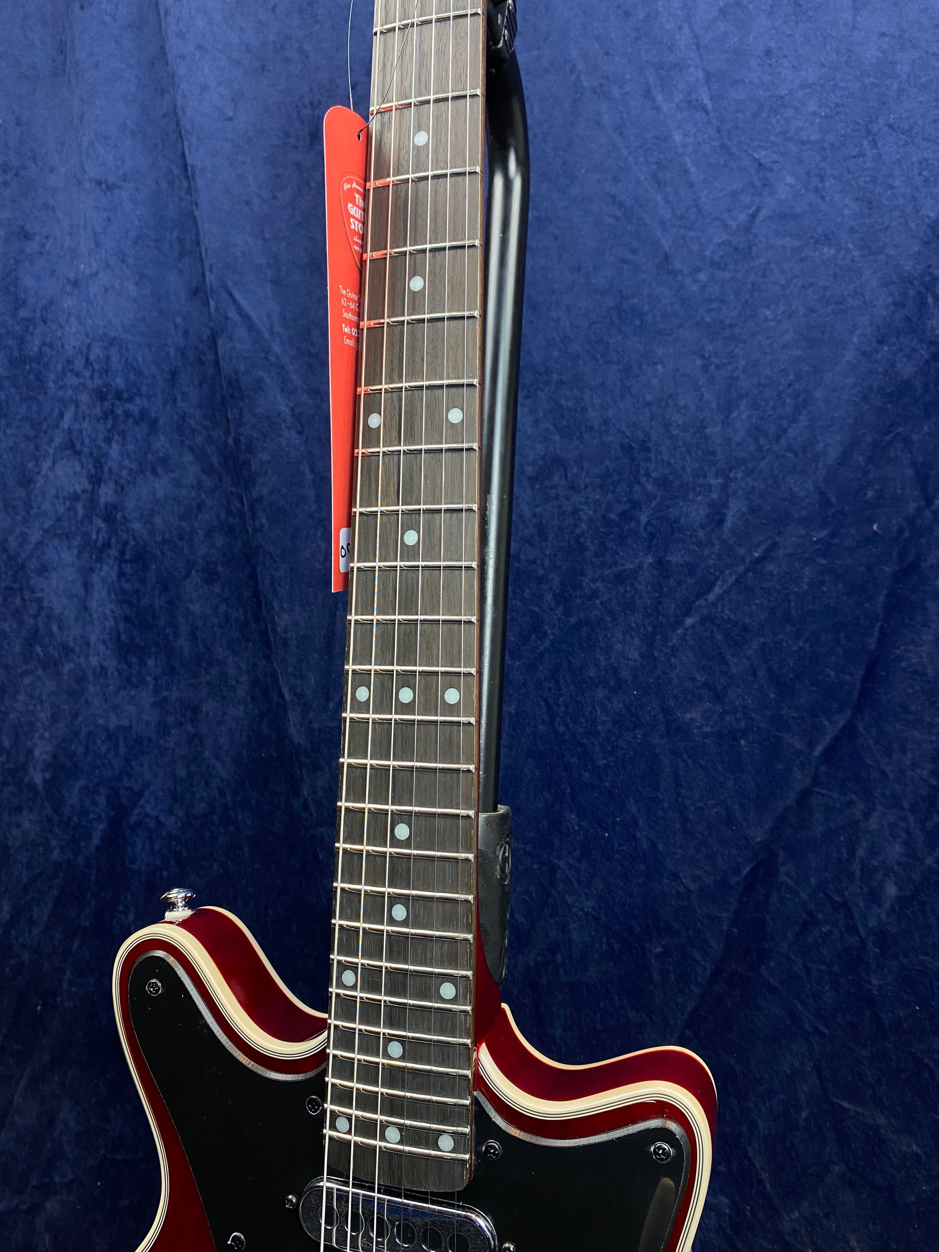 Red electric guitar with a dark wood fretboard, white dot inlays, and cream and black trim on the body. A red tag hangs from the neck.