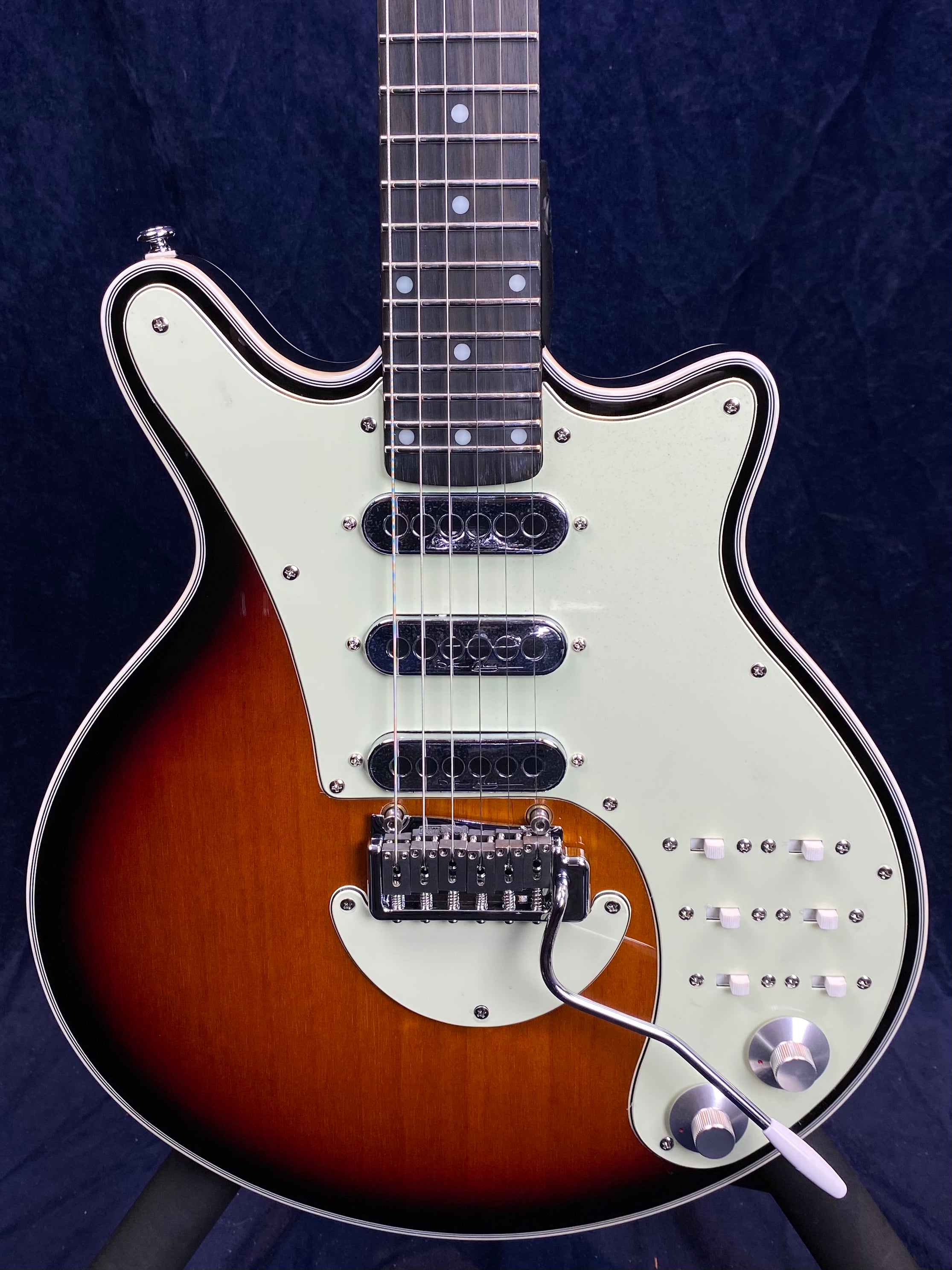 Sunburst electric guitar with three pickups, white pickguard, multiple toggle switches, two knobs, and a whammy bar on a contoured body.