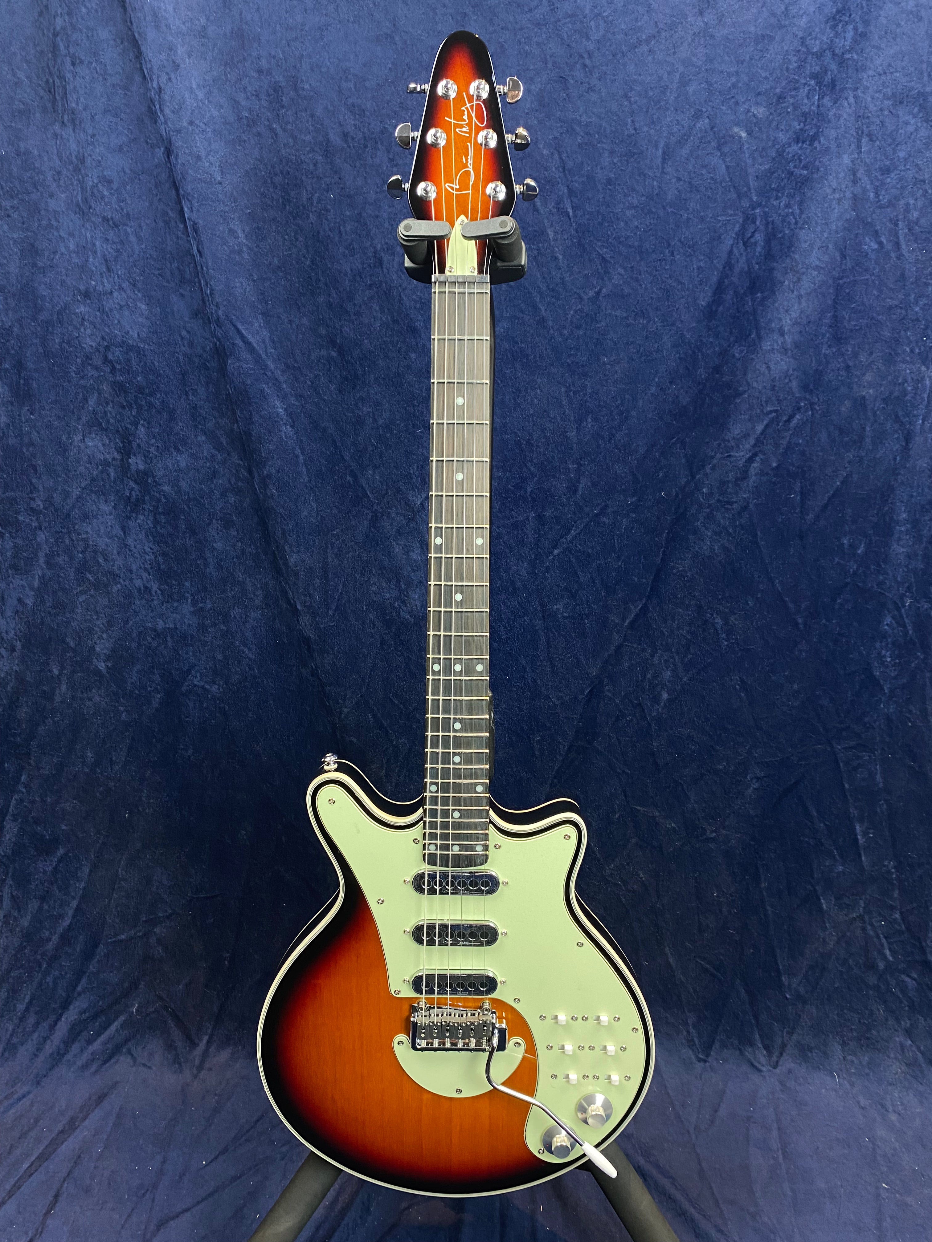 Electric guitar with sunburst finish, three pickups, white pickguard, chrome hardware, and tremolo arm, standing upright against a dark blue background.