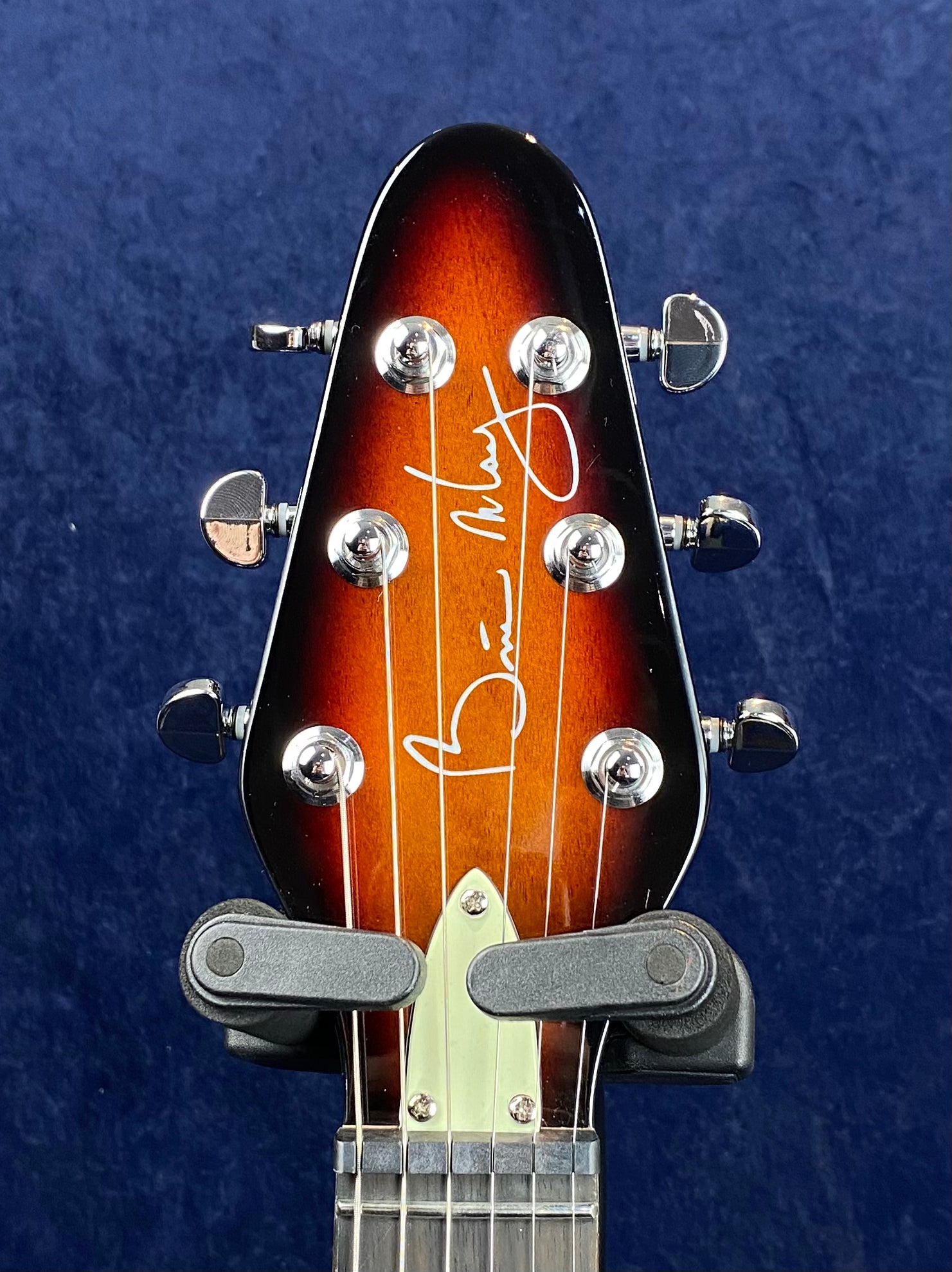 Sunburst electric guitar headstock with six tuning pegs, white signature reading "Brian May" across the front, and a dark blue background.