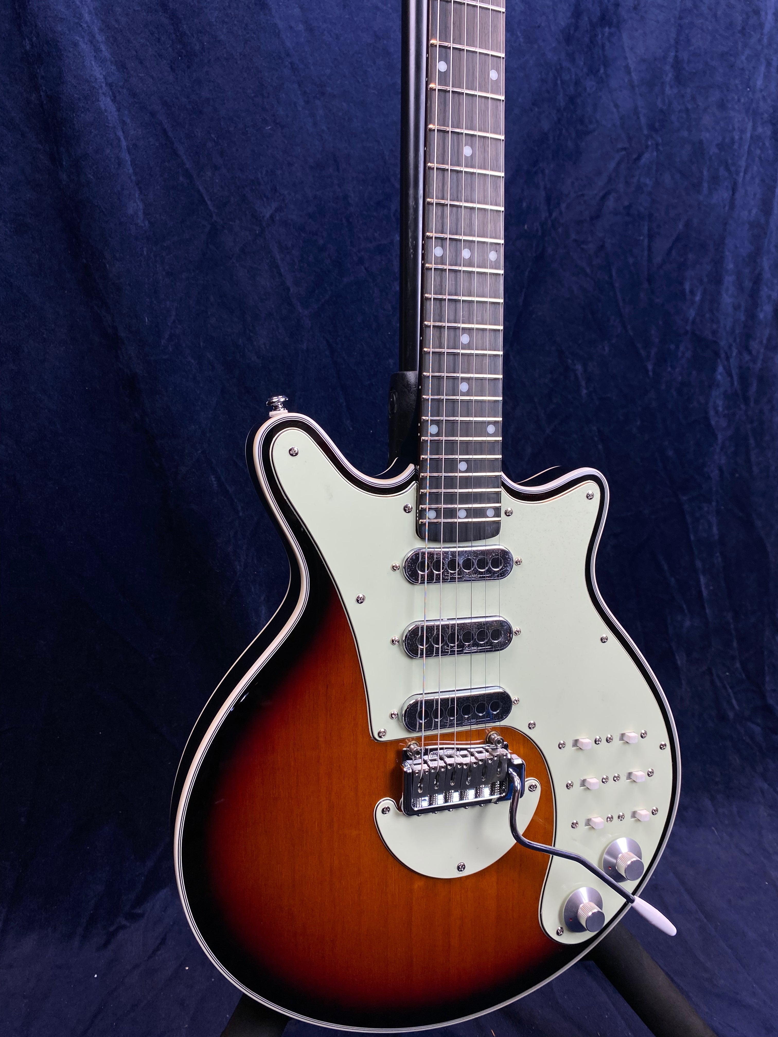 Sunburst electric guitar with three single-coil pickups, five switches, two knobs, a whammy bar, and a white pickguard against a dark background.