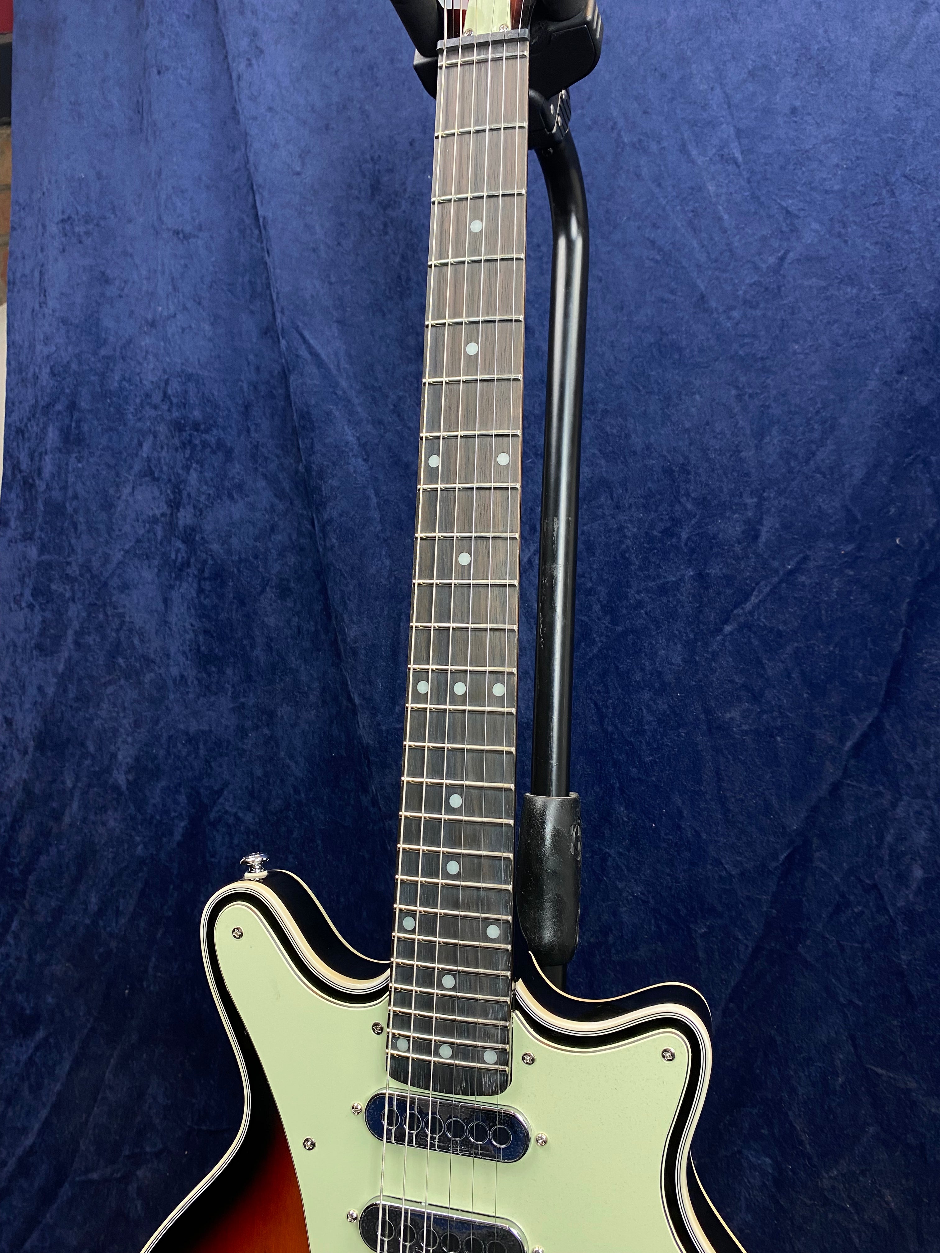 Electric guitar with a sunburst body, white pickguard, two pickups, and a dark wood fretboard with dot inlays, shown hanging on a stand.