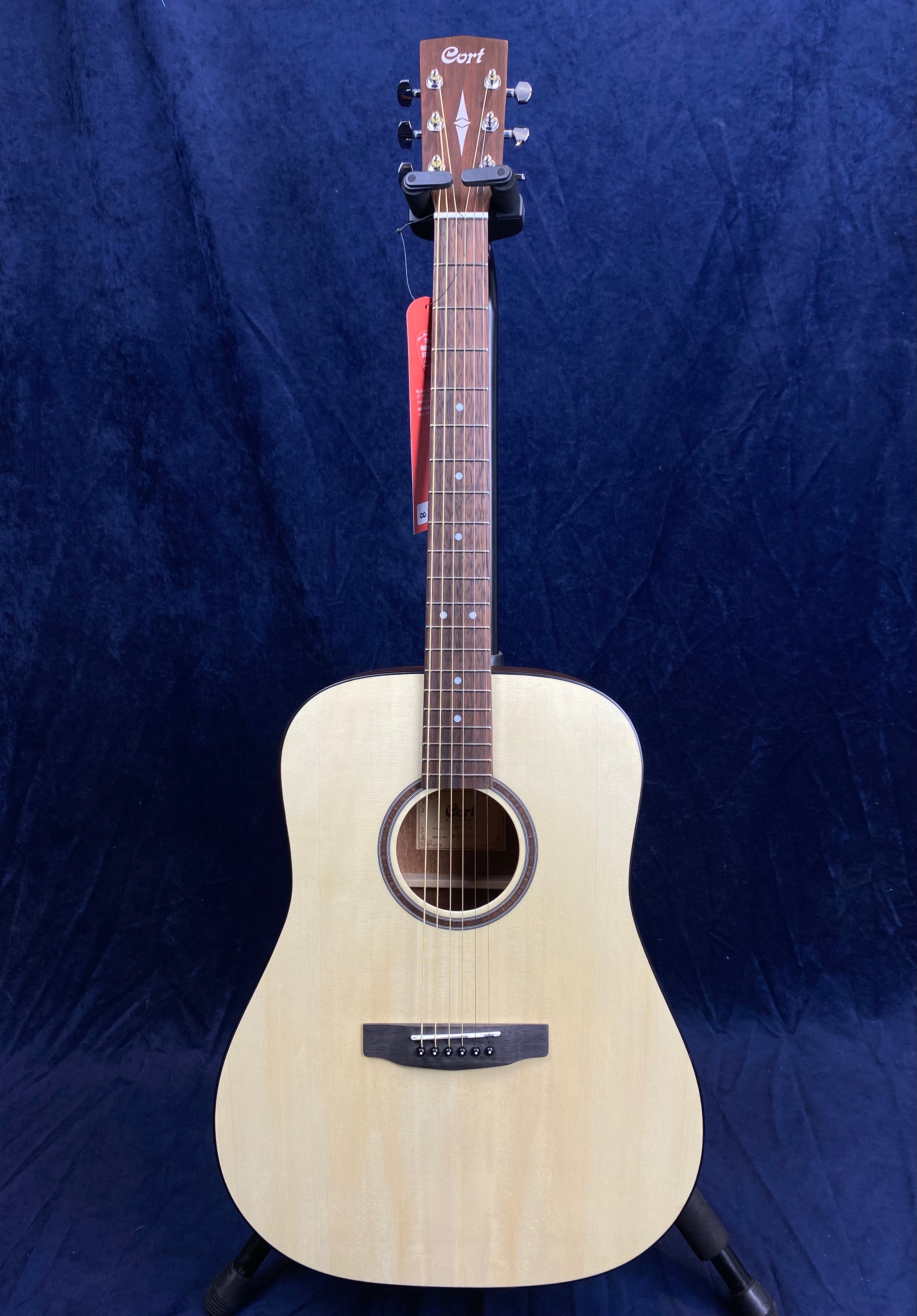 Natural wood finish Cort acoustic guitar with a rounded rectangular body, six strings, a brown fingerboard, and simple circular sound hole design.