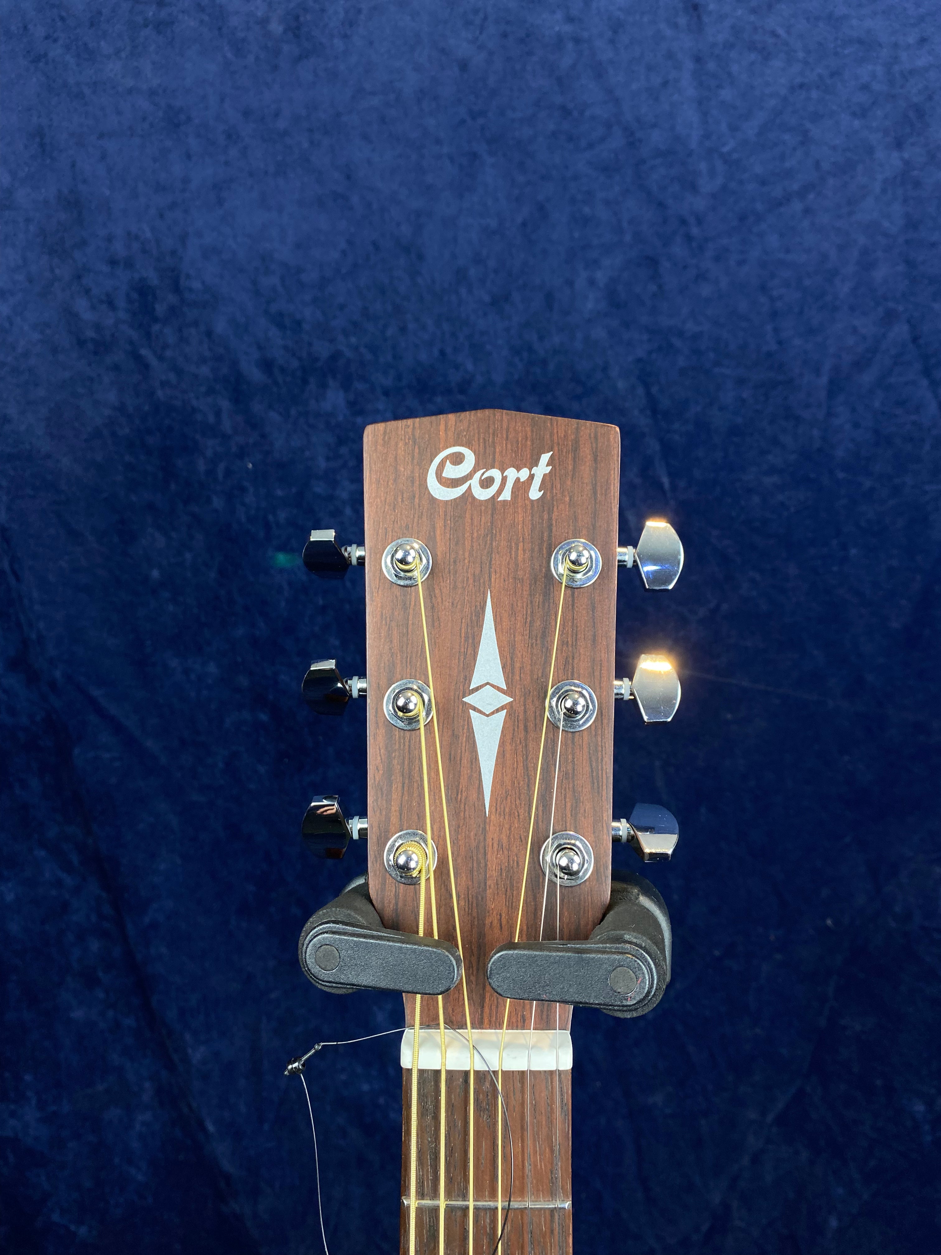 Headstock of a Cort acoustic guitar with six tuning pegs, a geometric inlay, and a dark wood finish against a dark blue background.