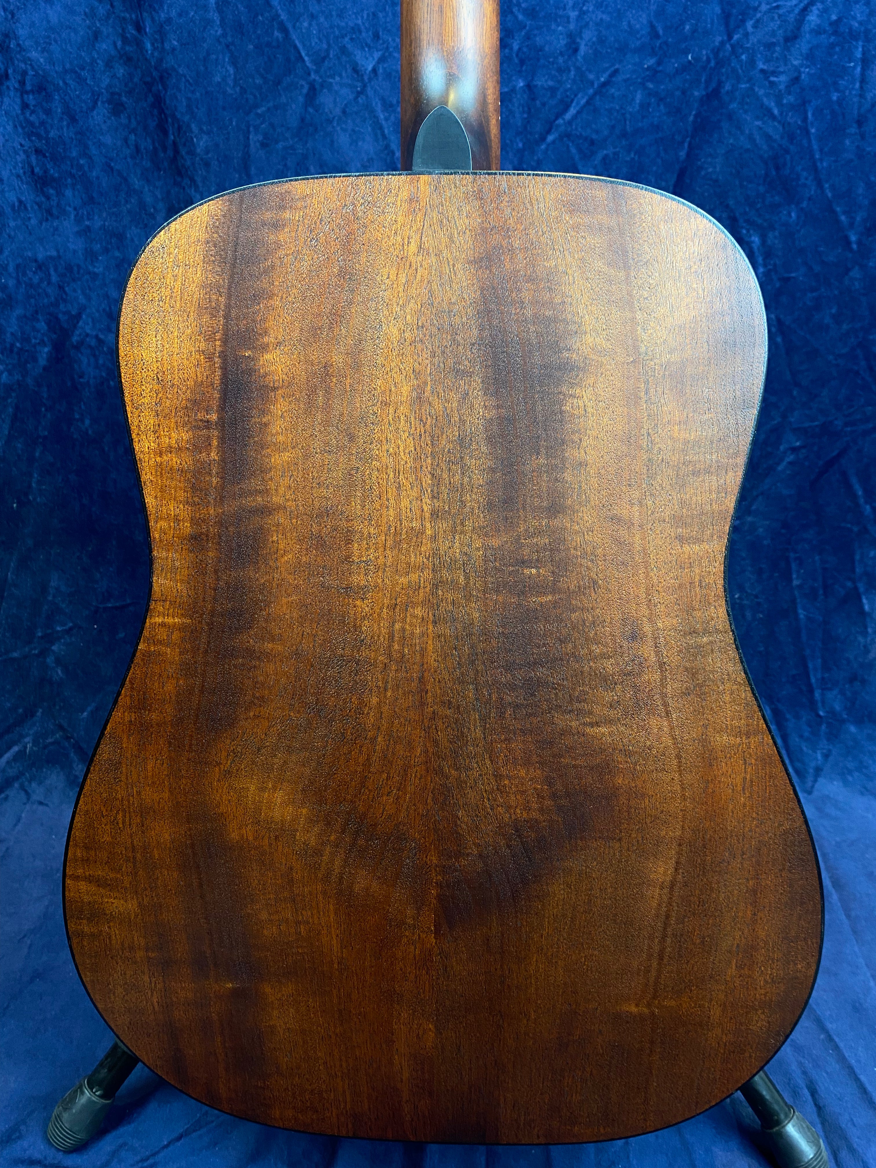 Back of an acoustic guitar showing smooth, dark brown wood grain with subtle lighter patterns, standing upright against a dark blue background.