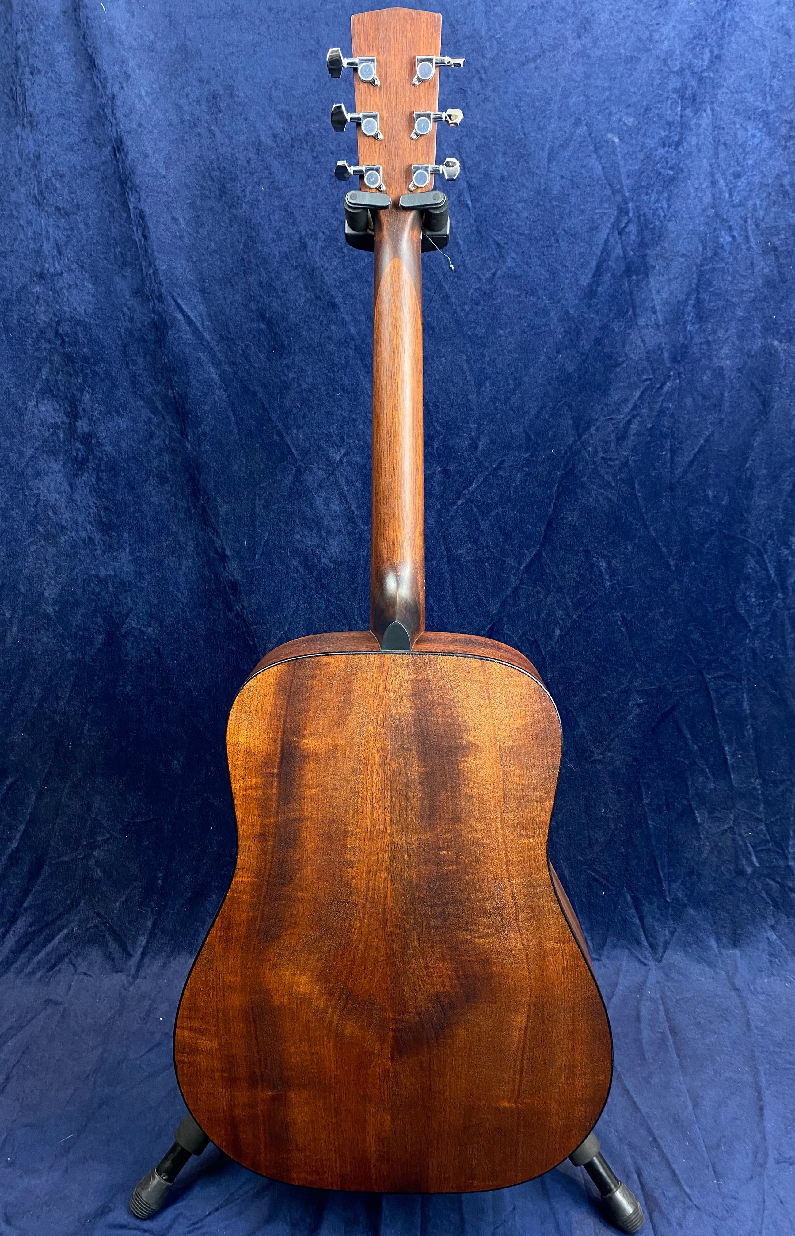 Acoustic guitar seen from the back, showing rich brown wood grain, six tuning pegs, and a curved dreadnought body against a dark blue background.