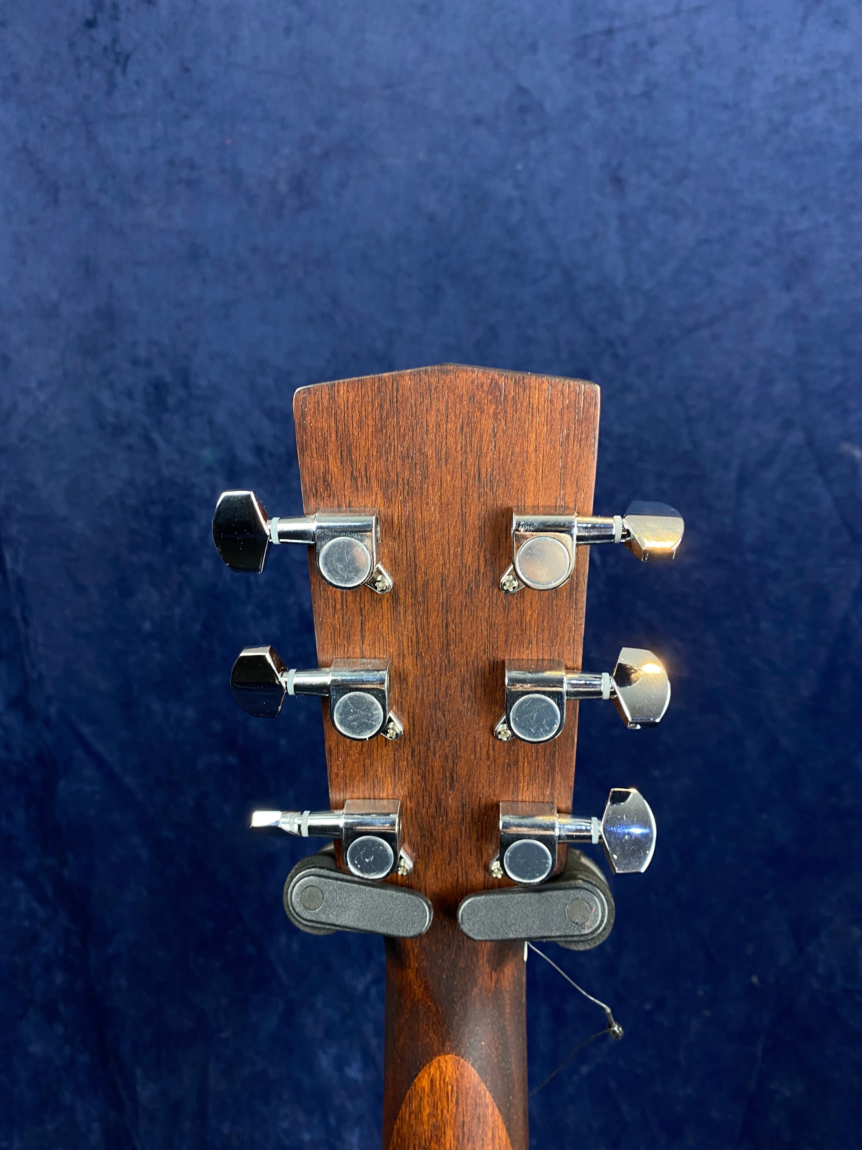 Rear view of an acoustic guitar headstock with six chrome tuning pegs and a dark wooden finish against a blue background.