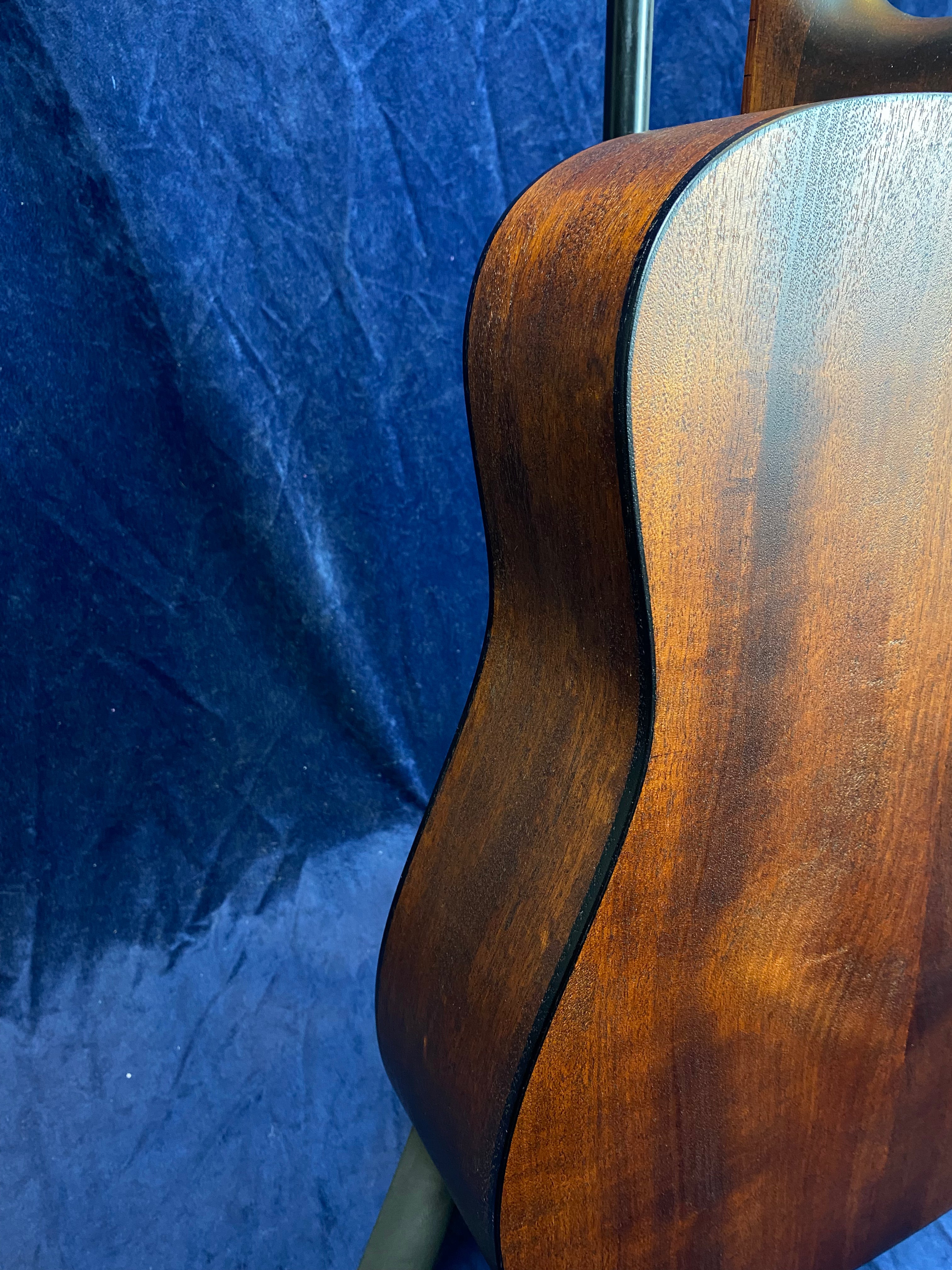 Curved wooden side and back of an acoustic guitar, showing rich brown grain and smooth finish, against a textured dark blue background.