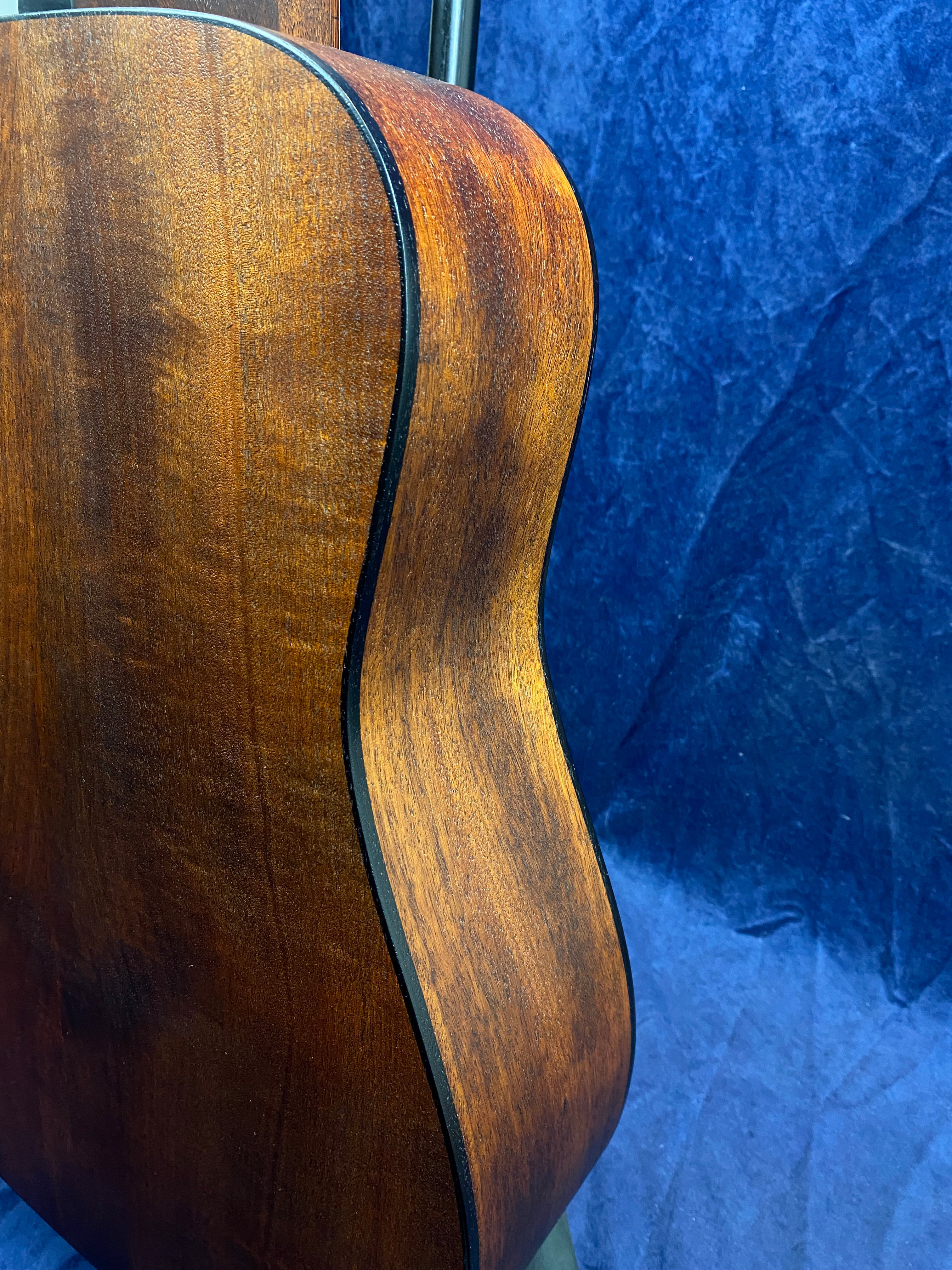Glossy, rich brown wood side of an acoustic guitar with black binding, set against a deep blue textured background.
