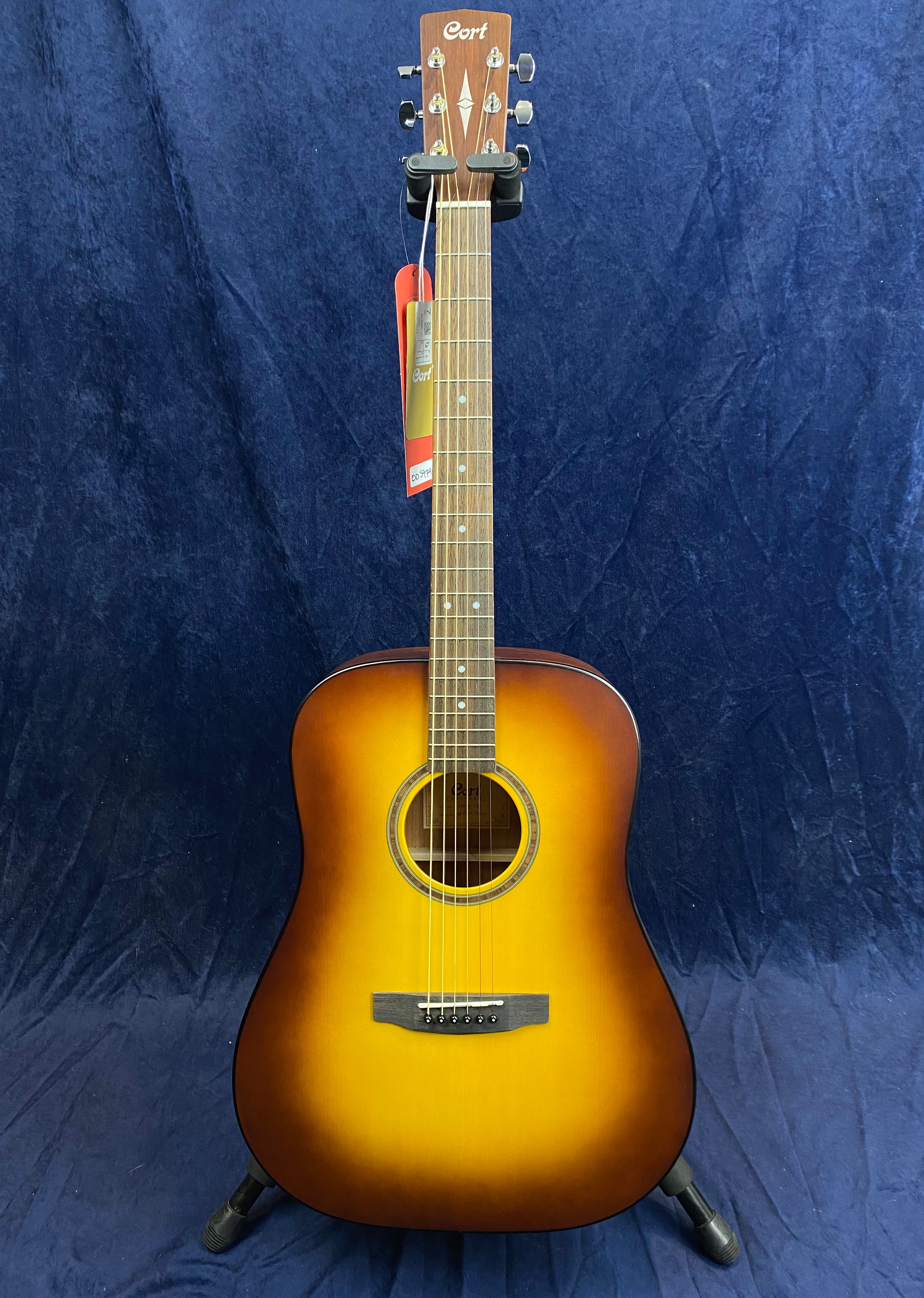 Sunburst Cort acoustic guitar with six strings, circular sound hole, and tags hanging from the headstock, standing upright on a floor stand.