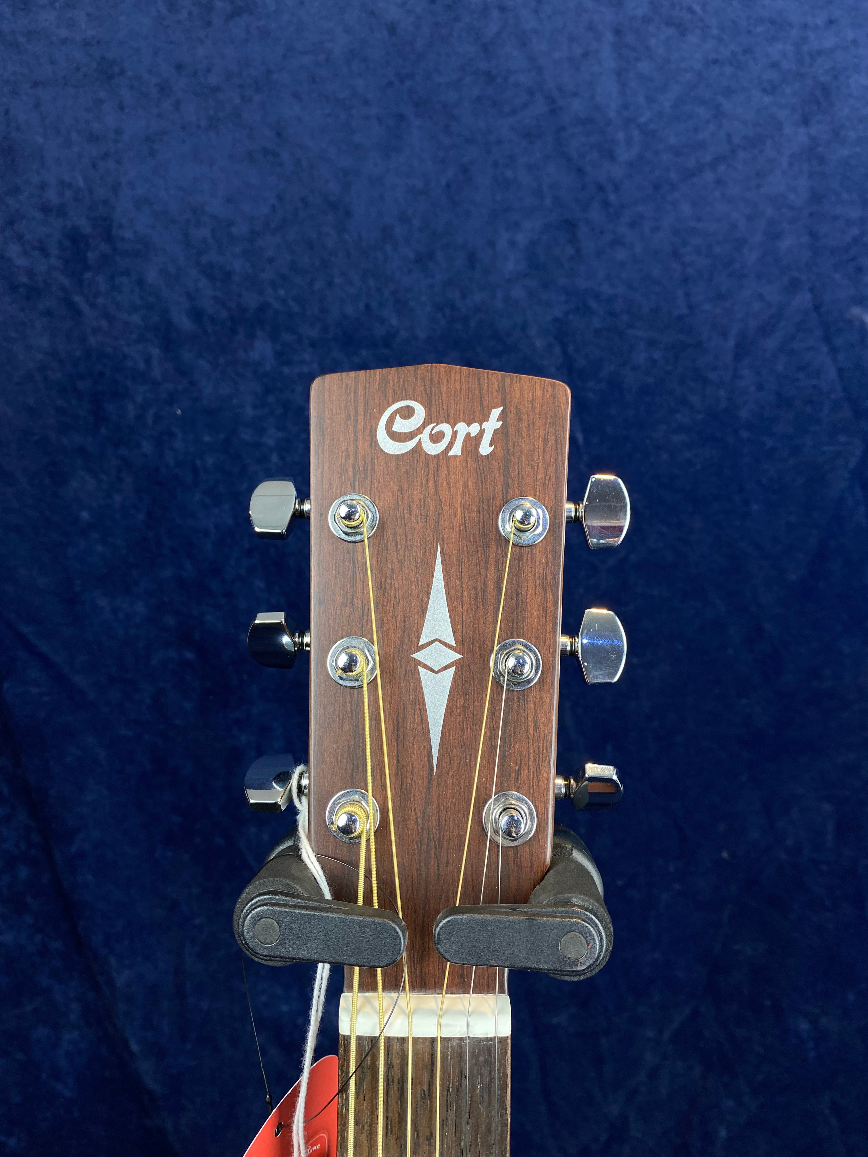 The headstock of a Cort acoustic guitar with six tuning pegs, wood grain finish, and a silver geometric inlay below the Cort logo.