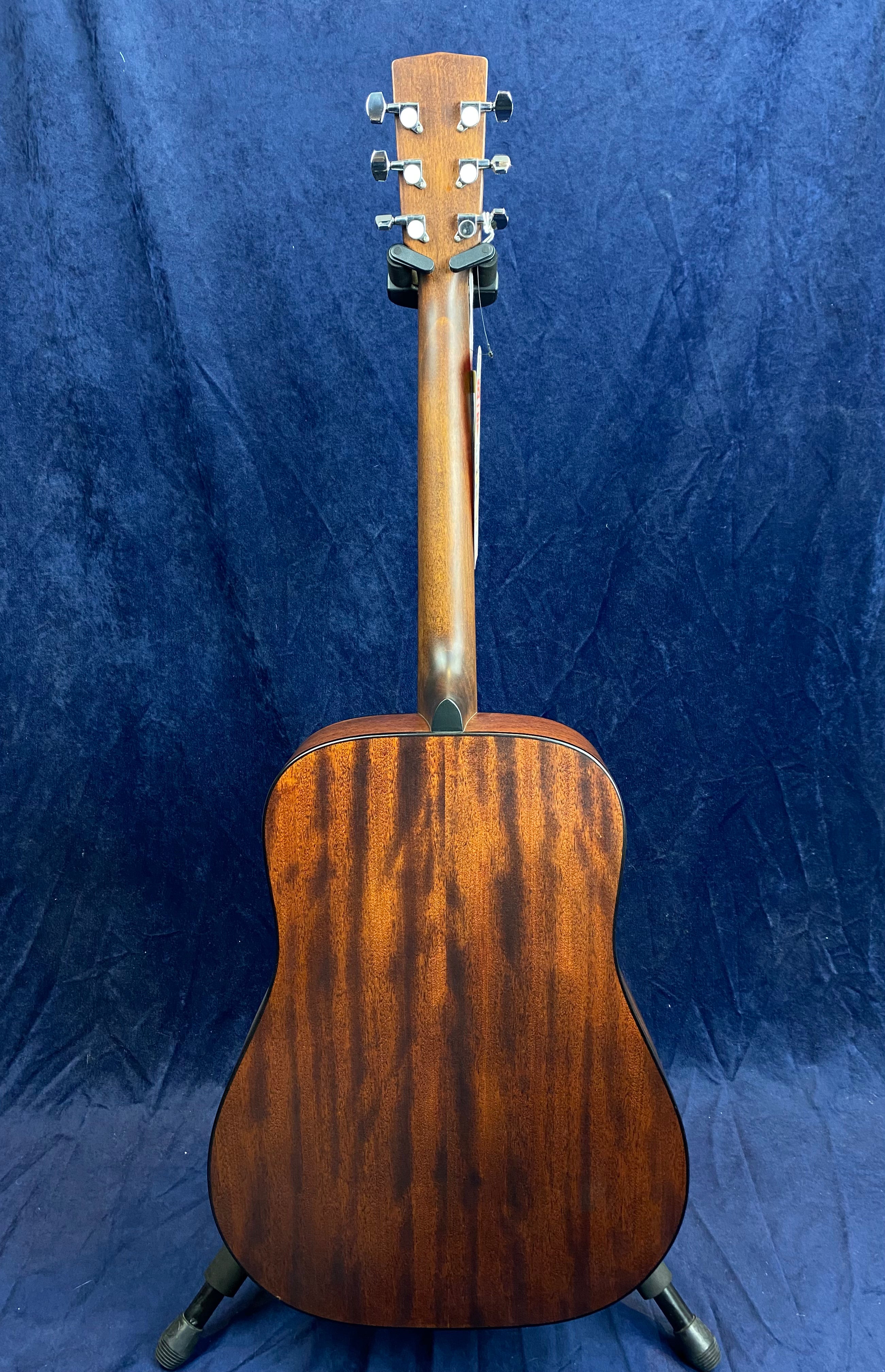 Mahogany acoustic guitar shown from the back, displaying a rich brown wood grain, silver tuning pegs, and a smooth, curved body on a stand.