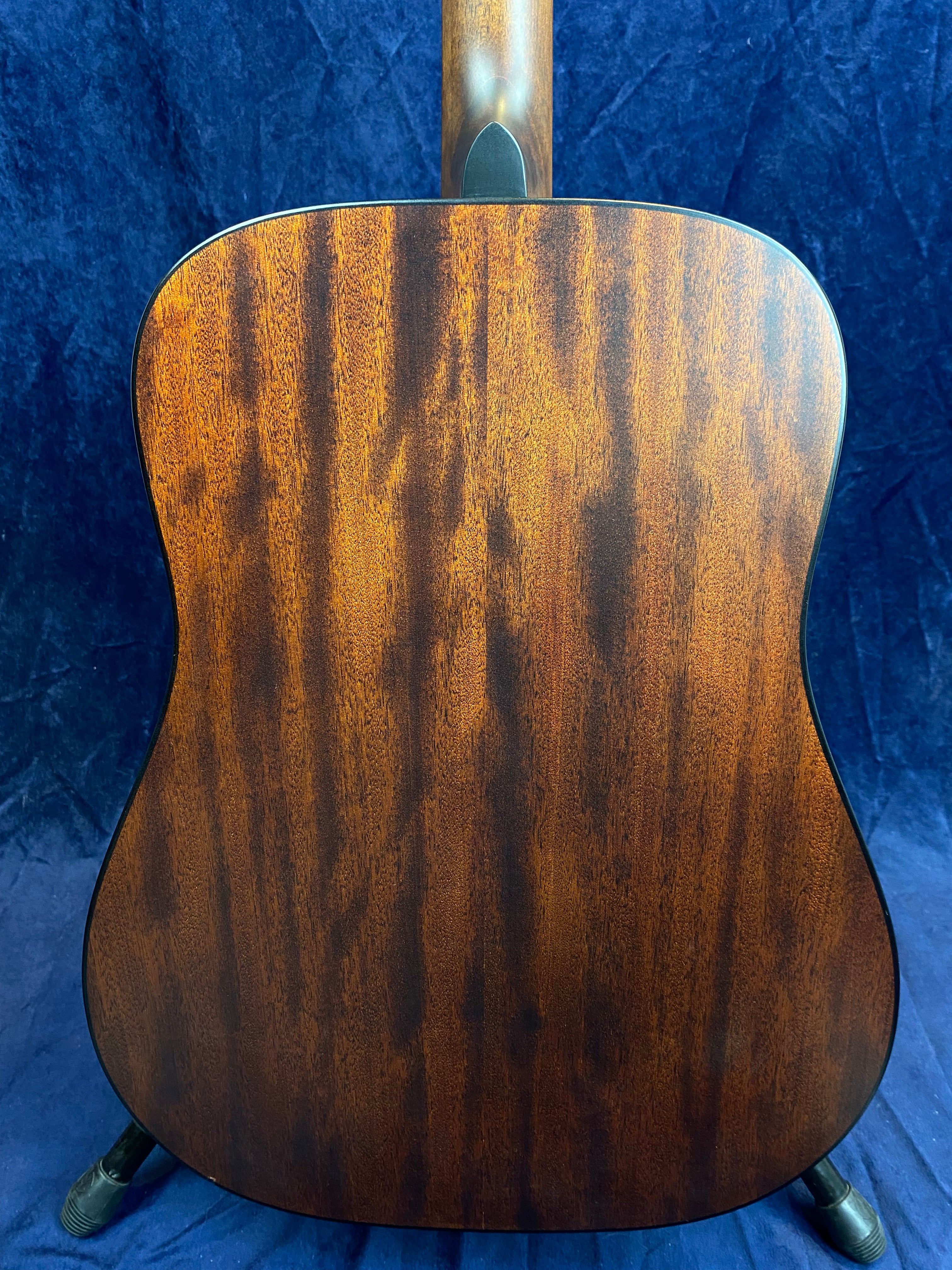 Back view of an acoustic guitar showing dark brown wood with vertical grain patterns and a smooth, glossy finish against a dark blue background.