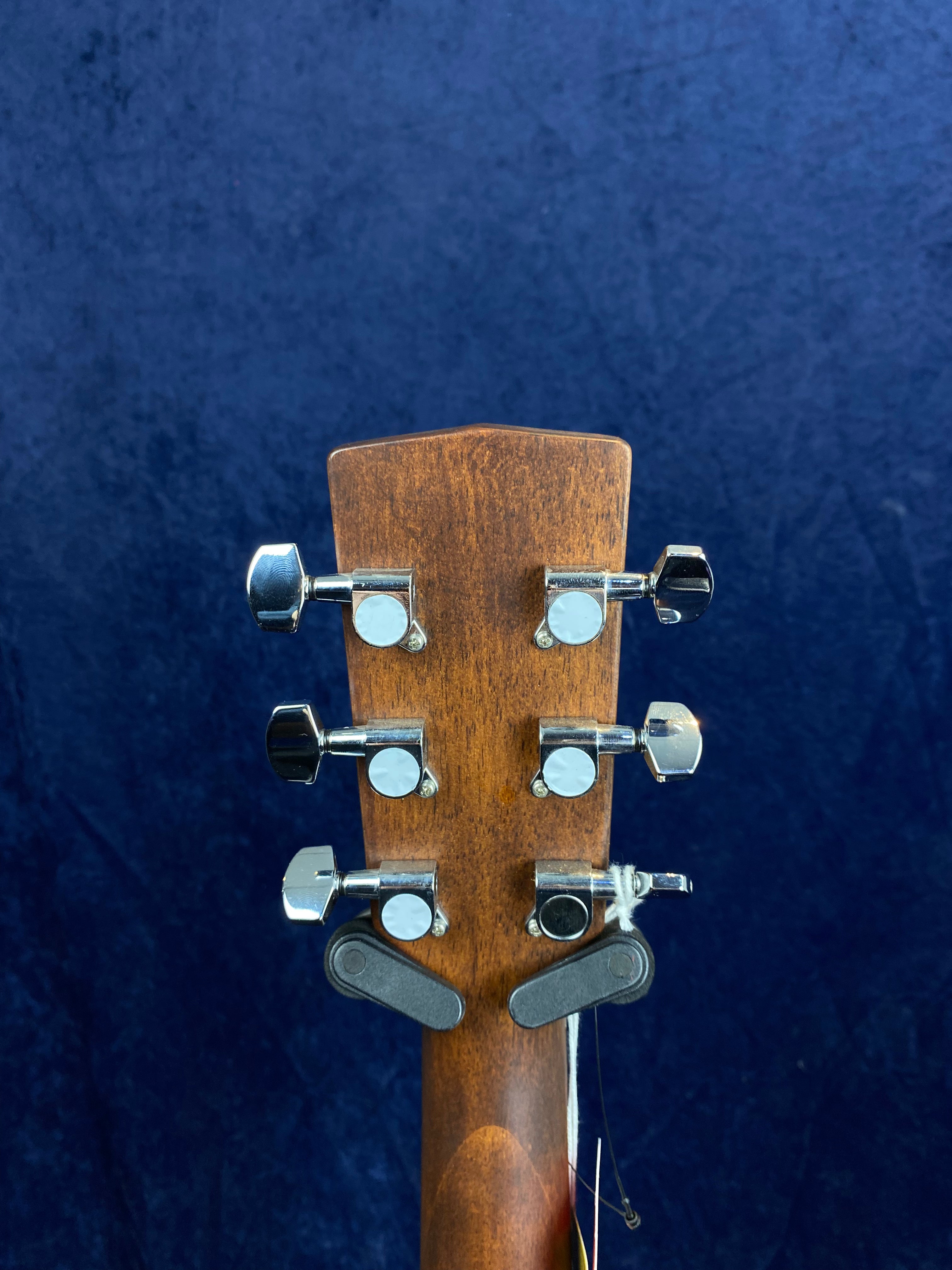 Back of an acoustic guitar headstock showing six chrome tuning pegs on a brown wood finish with a dark blue background.