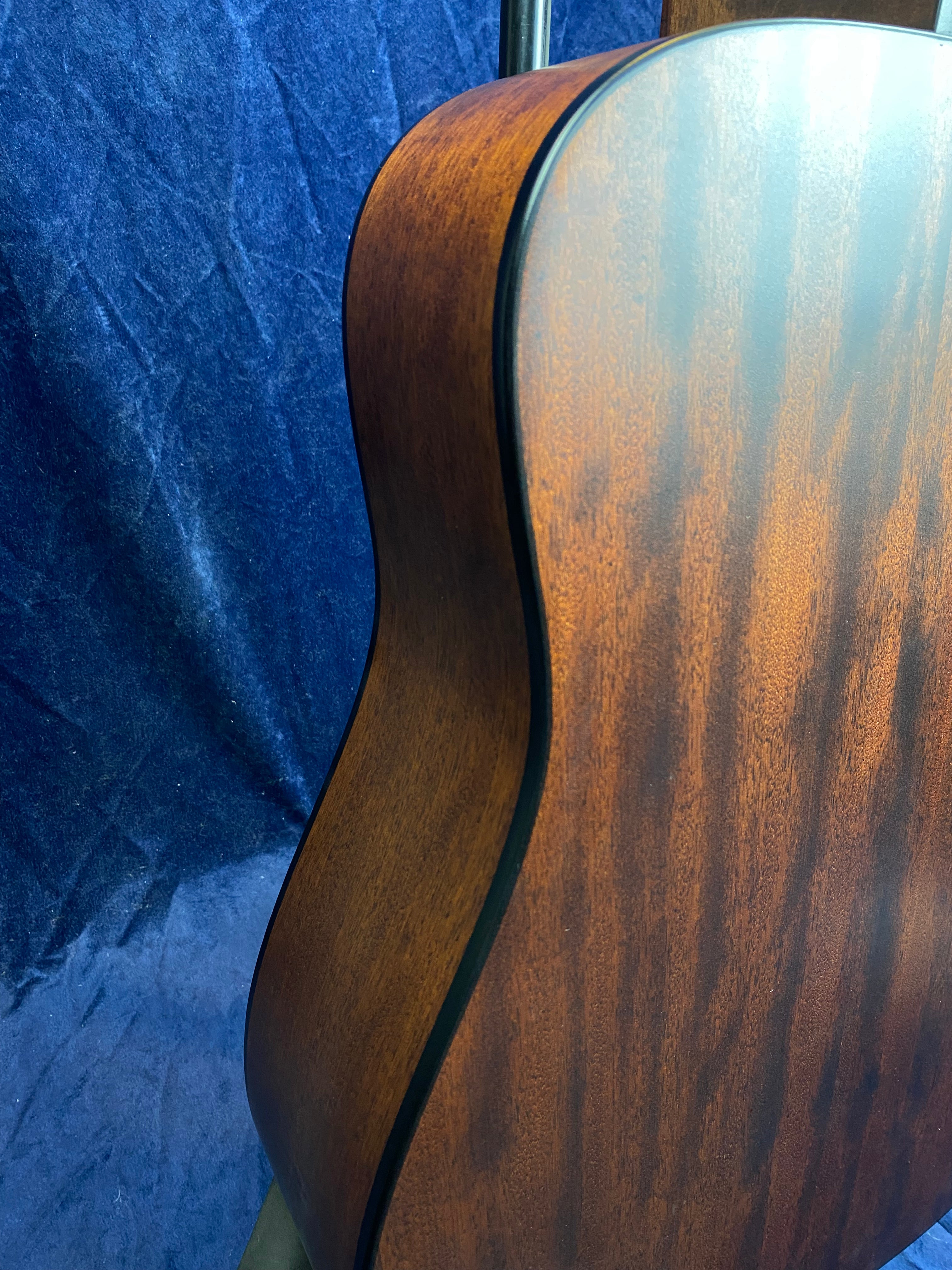 Curved side and back view of an acoustic guitar, showing smooth, rich brown wood with dark grain patterns against a blue textured background.
