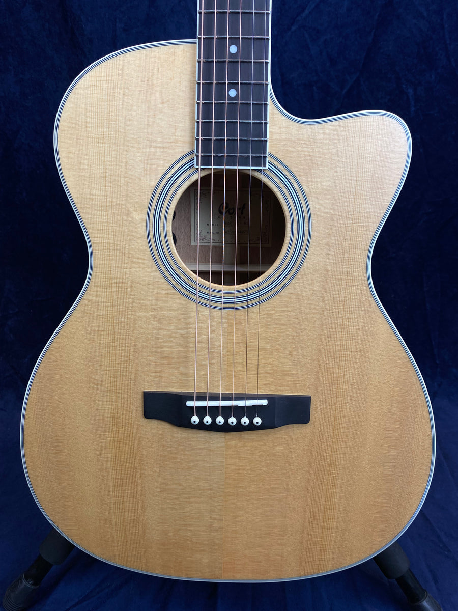 Light wood acoustic guitar with a cutaway body, six strings, dark bridge with white pins, and circular sound hole outlined by multiple rings.