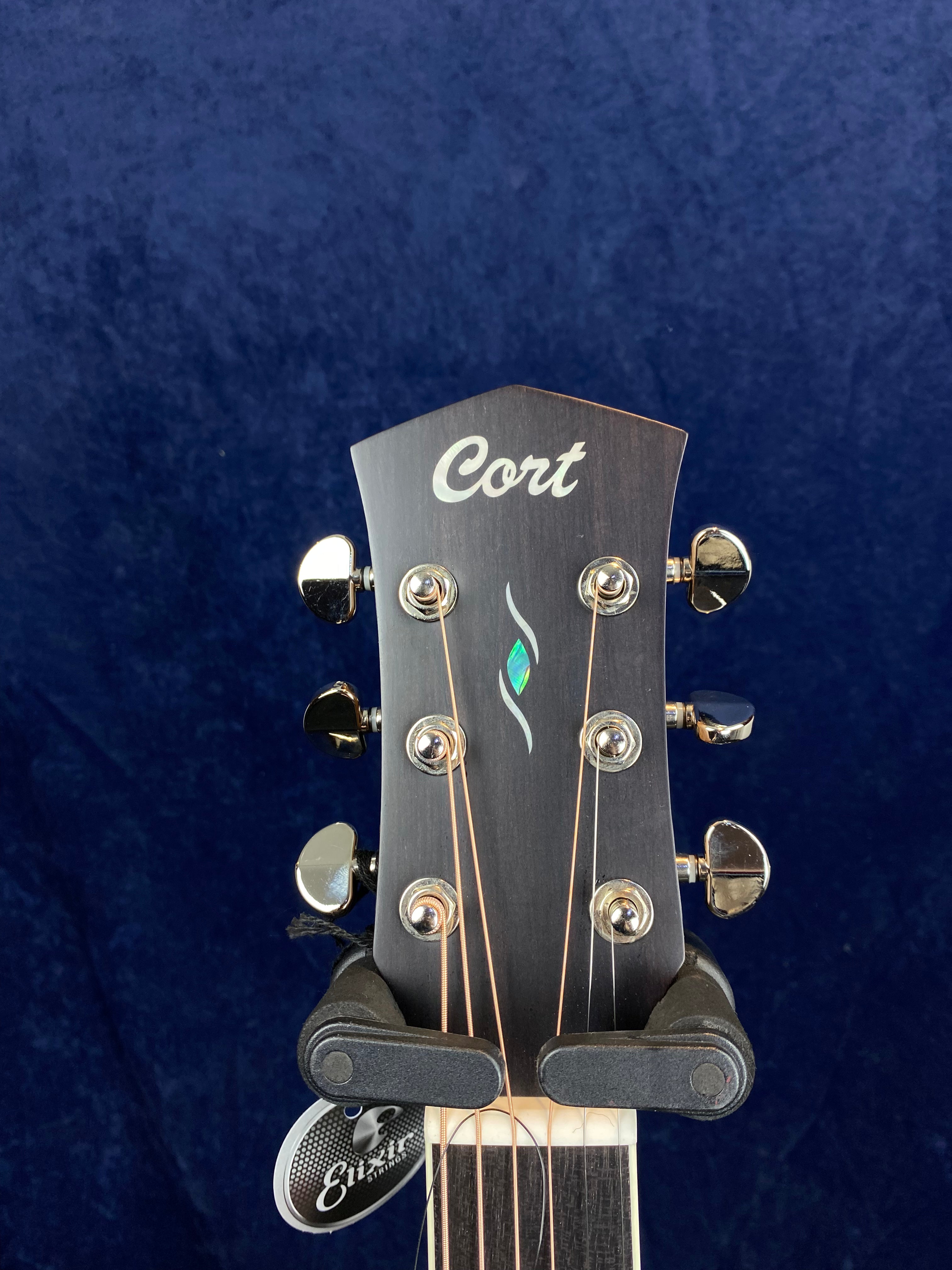 Cort acoustic guitar headstock with six tuning pegs, a small abalone inlay, and strings attached, set against a dark blue background.