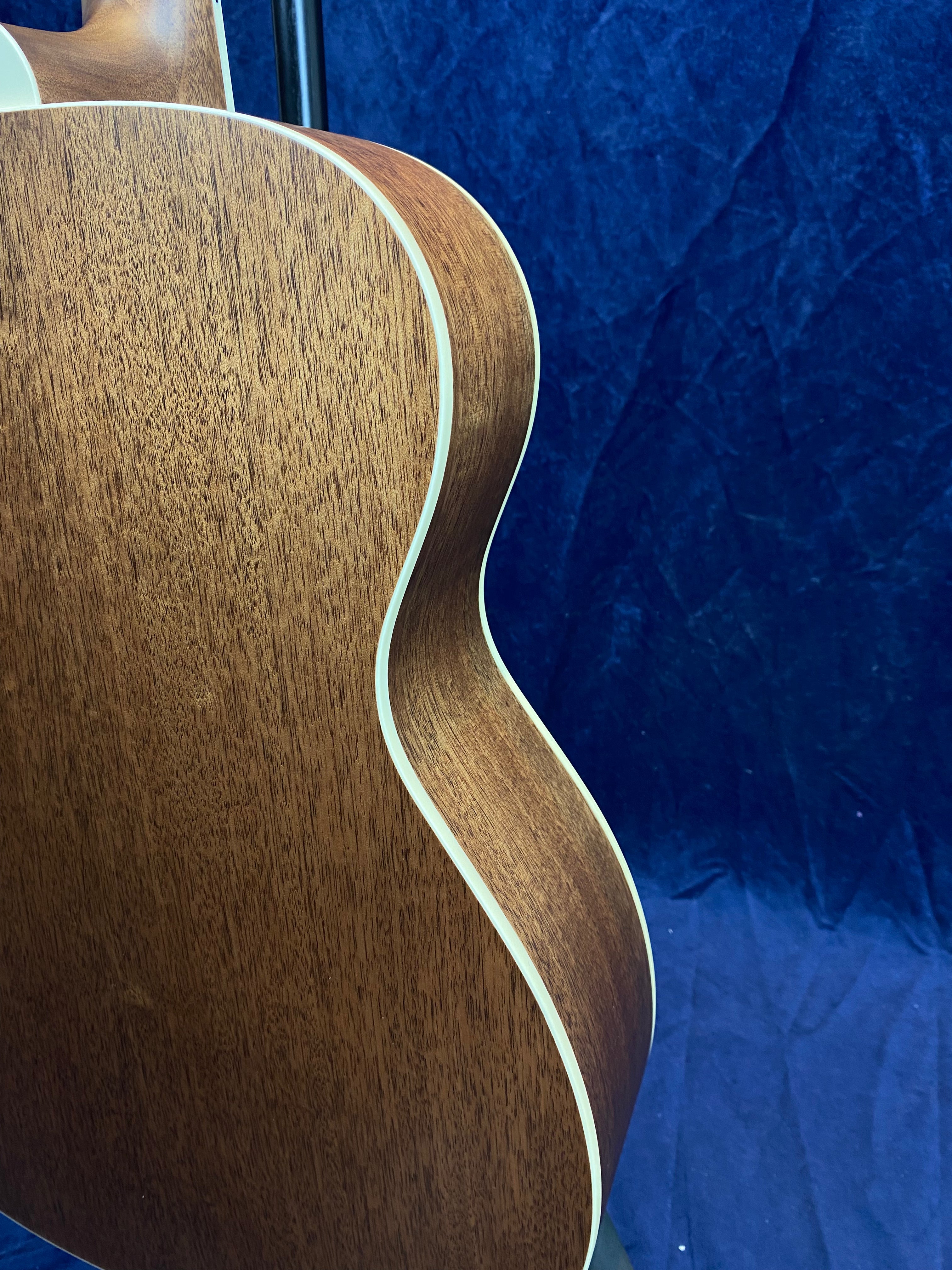Curved mahogany wood side and back of an acoustic guitar with cream binding, set against a textured dark blue background.