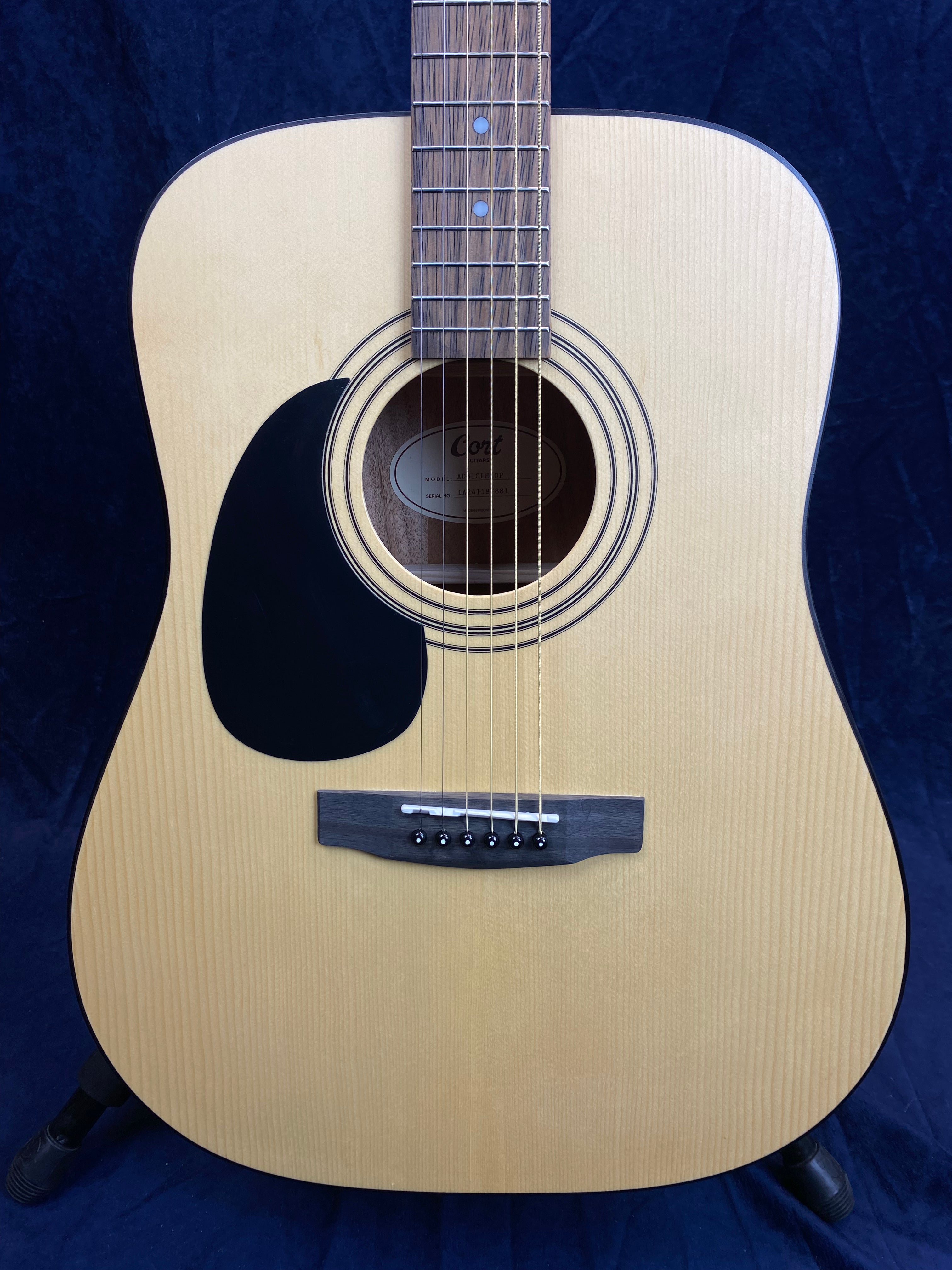 Light wood acoustic guitar with black pickguard, round sound hole, six strings, and a simple bridge, shown against a dark background.