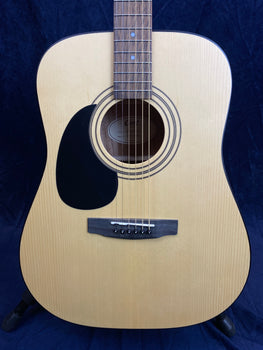 Light wood acoustic guitar with black pickguard, round sound hole, six strings, and a simple bridge, shown against a dark background.