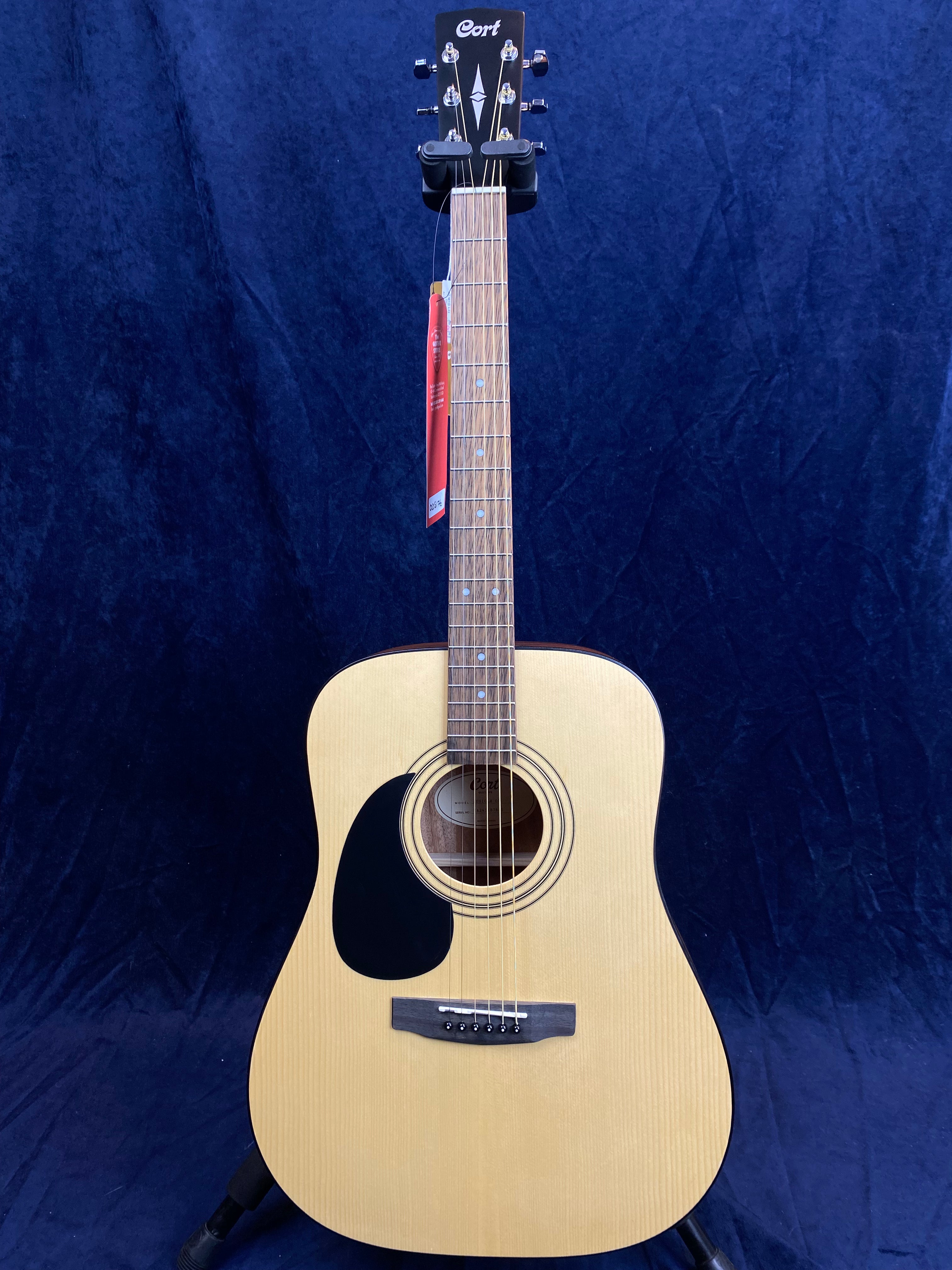 Natural wood finish acoustic guitar with a black pickguard, six strings, and a red tag hanging from the neck, standing against a dark blue background.