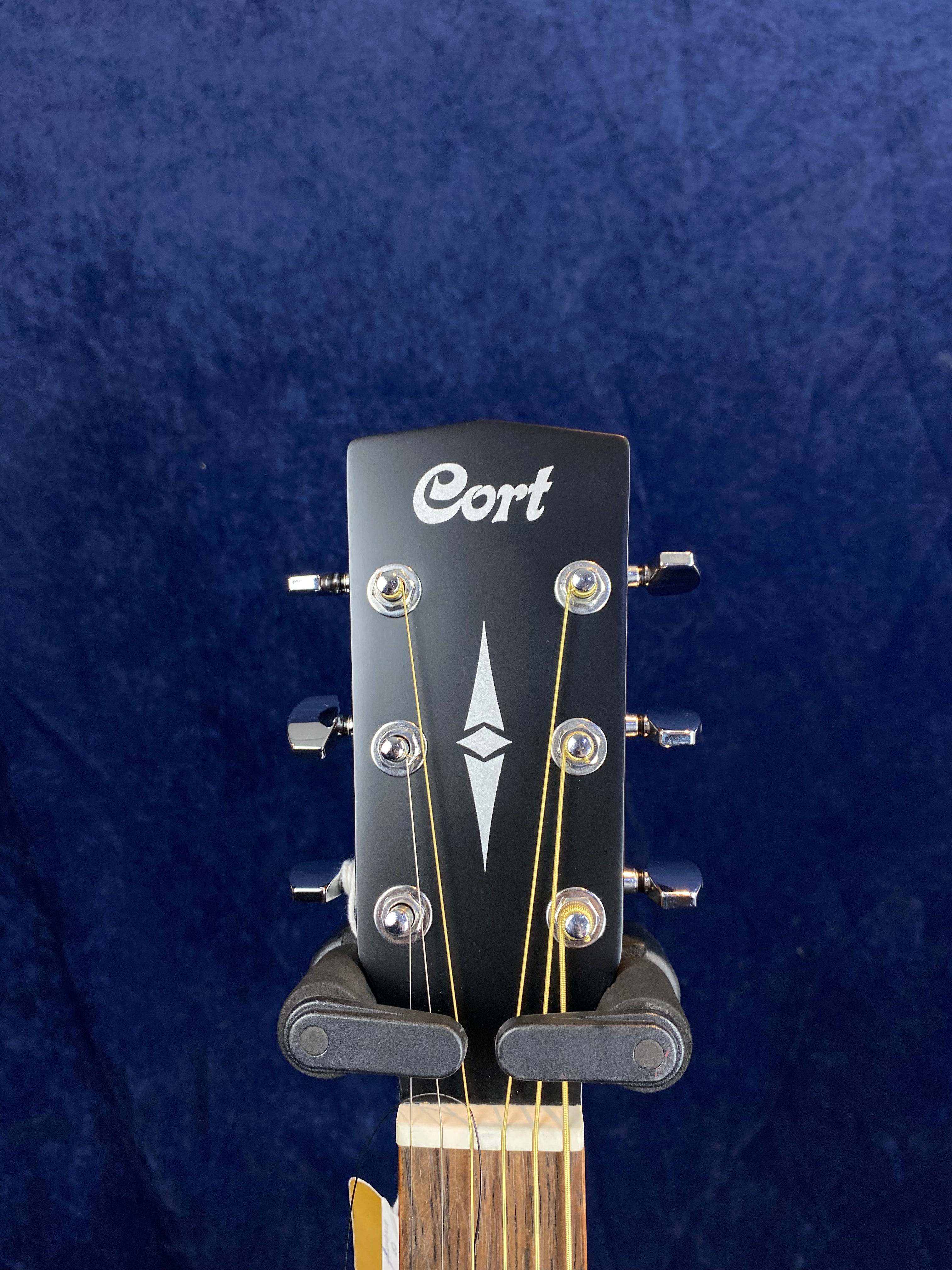 Black headstock of a Cort acoustic guitar with six tuning pegs, silver diamond inlay, and visible strings against a dark blue background.