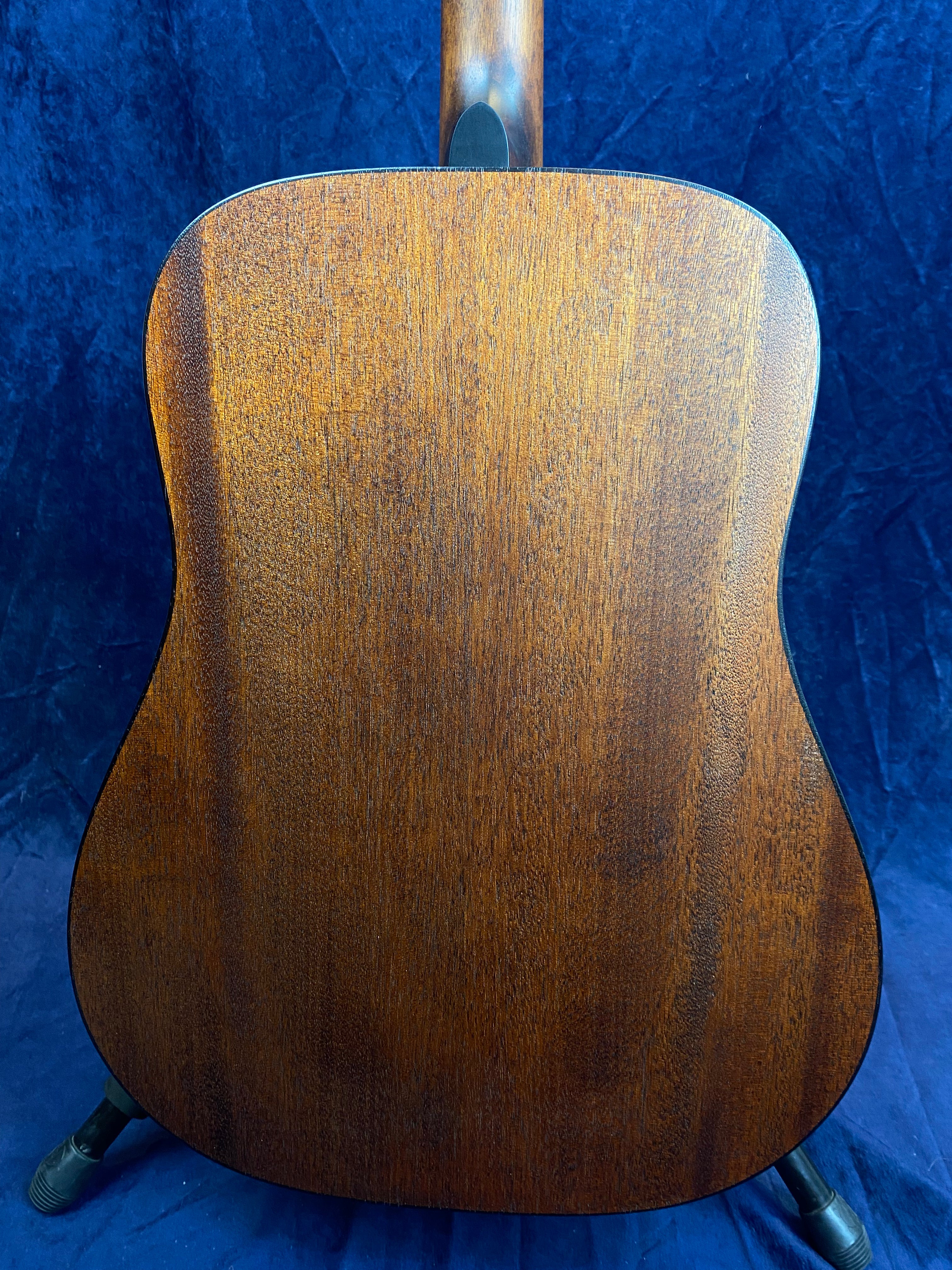 Acoustic guitar back shown, wooden surface with a rich brown color and natural grain patterns, blue fabric background, guitar stands visible at bottom.