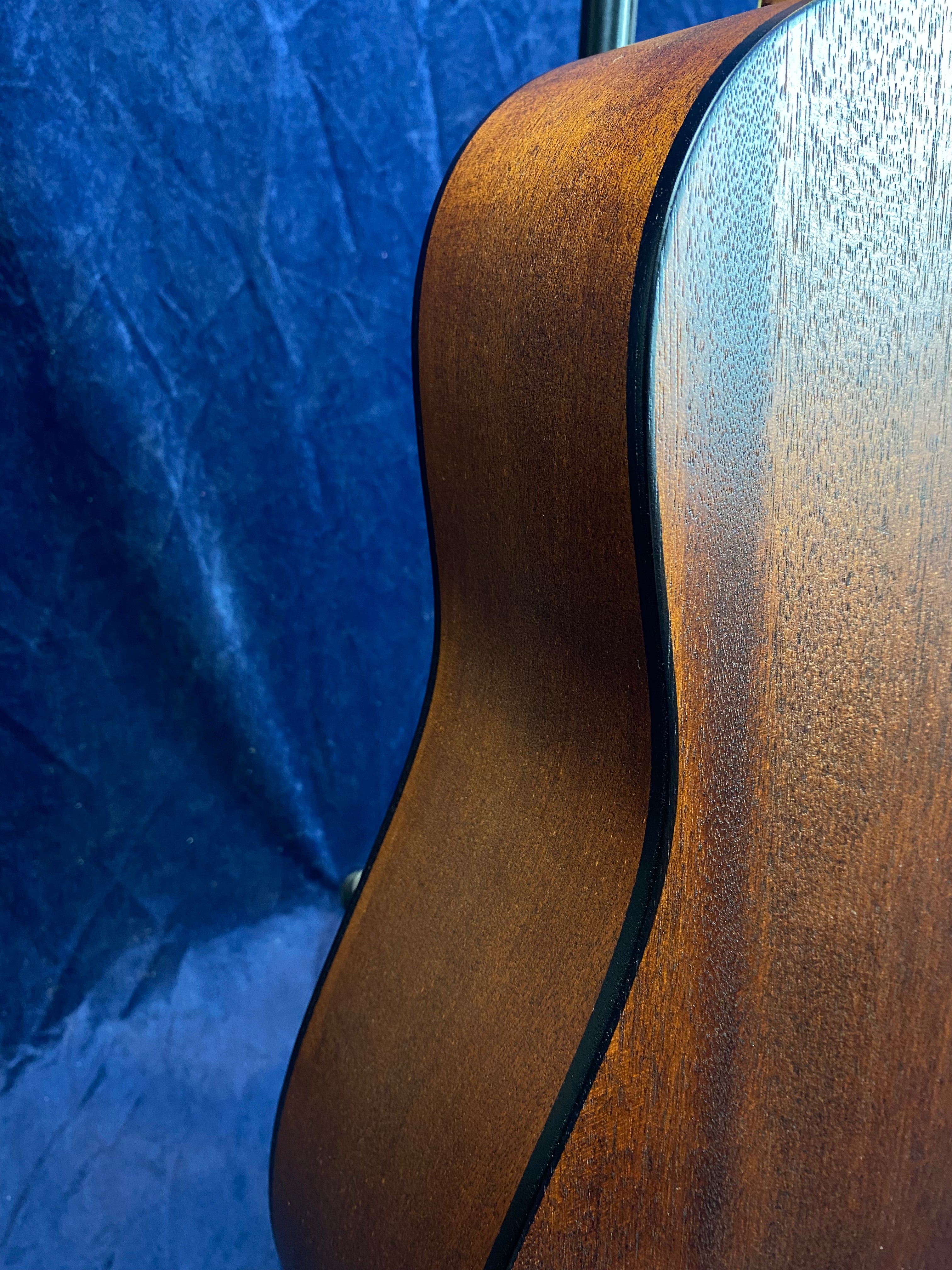 Curved side view of a brown acoustic guitar body with visible wood grain and black binding against a textured blue background.