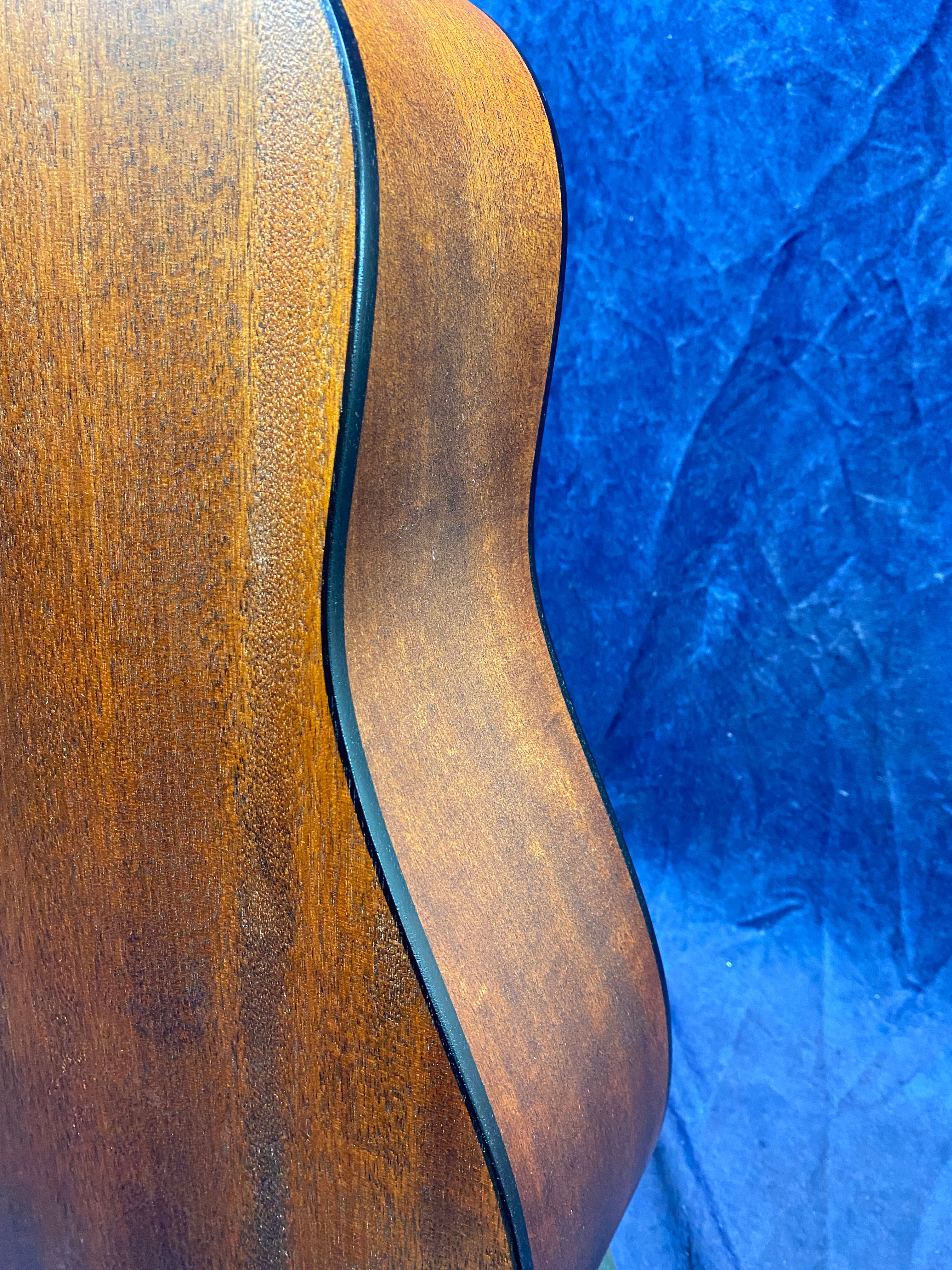 Curved side view of an acoustic guitar showing smooth, brown wood grain and black edge binding against a textured blue background.