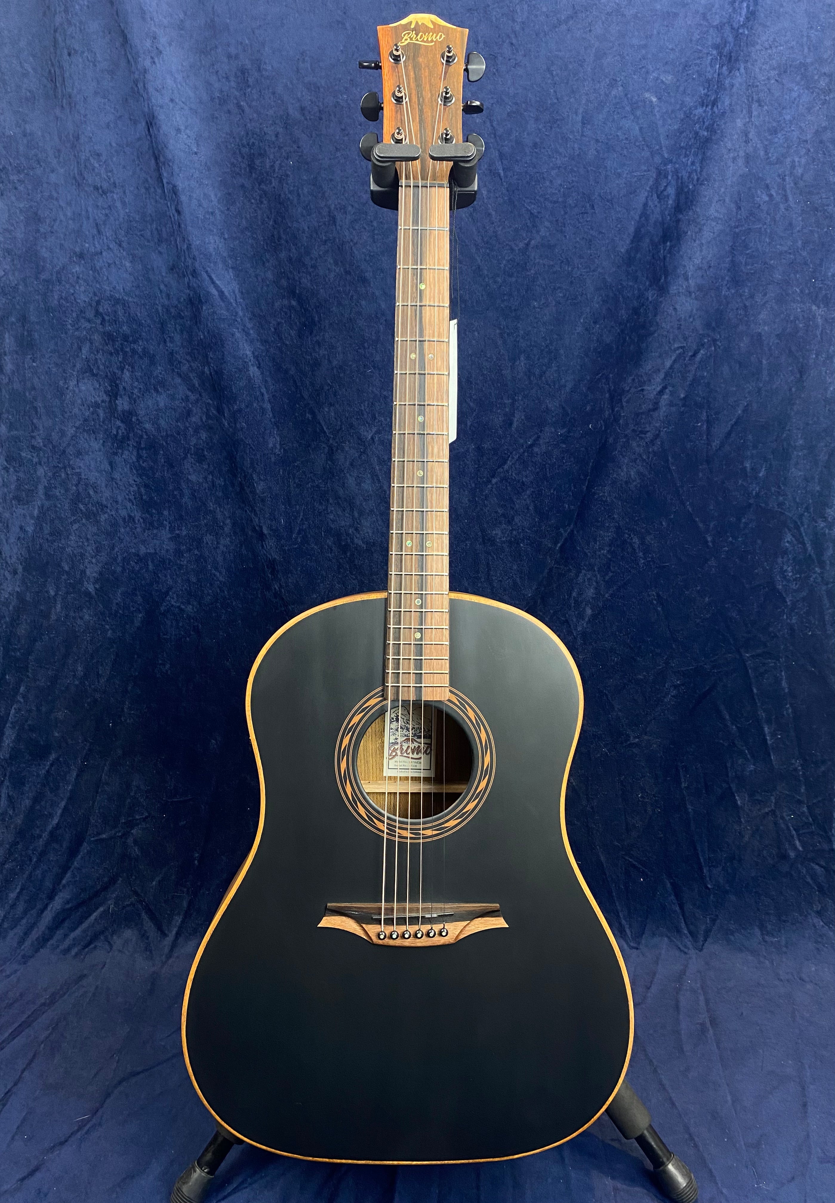 Black acoustic guitar with a natural wood neck and headstock, lighter wood trim, six strings, and decorative soundhole inlay, standing upright on a stand.