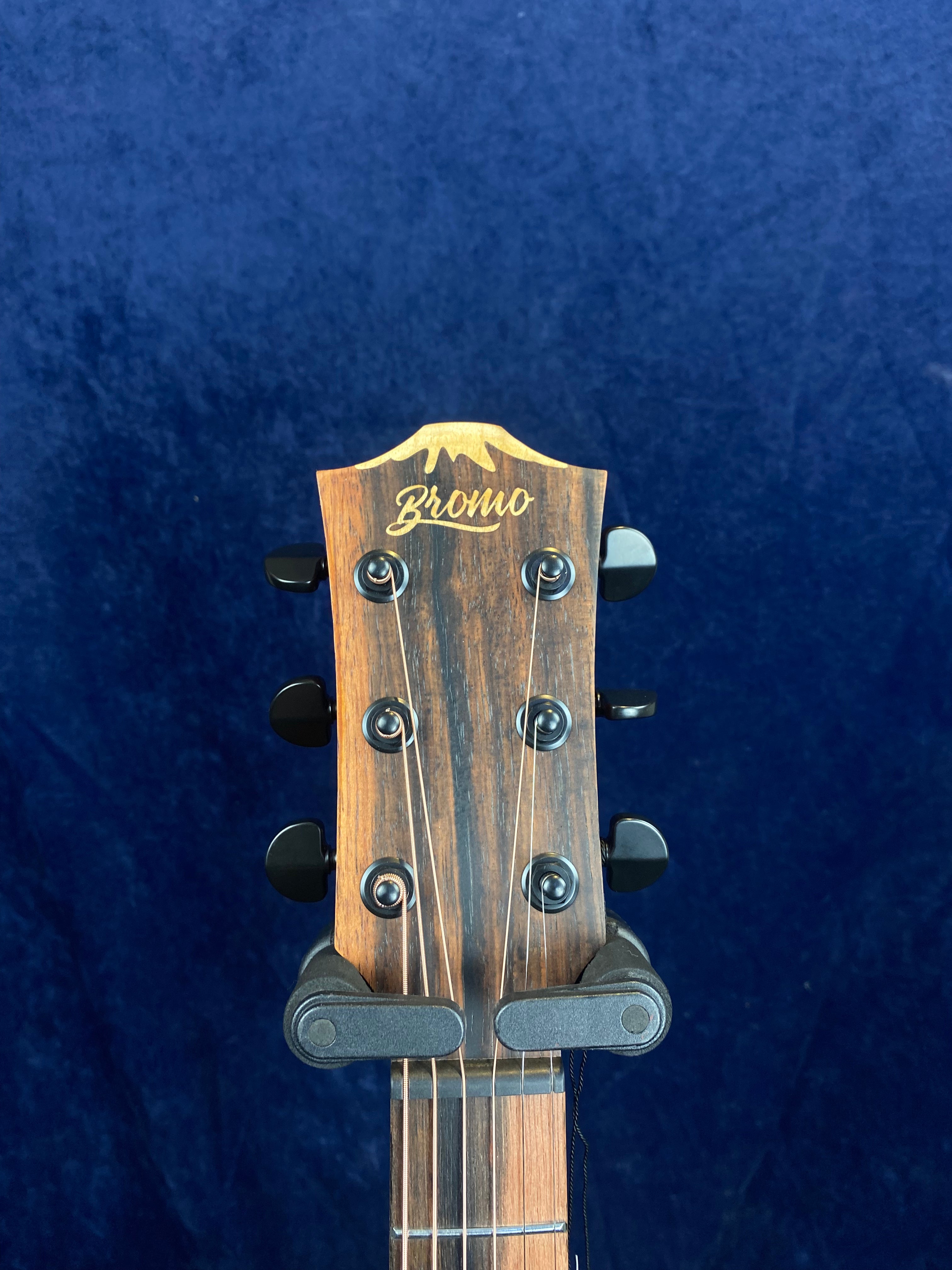 The headstock of a Bromo acoustic guitar with six tuning pegs, a wood finish, and a guitar capo clamped just below the headstock.