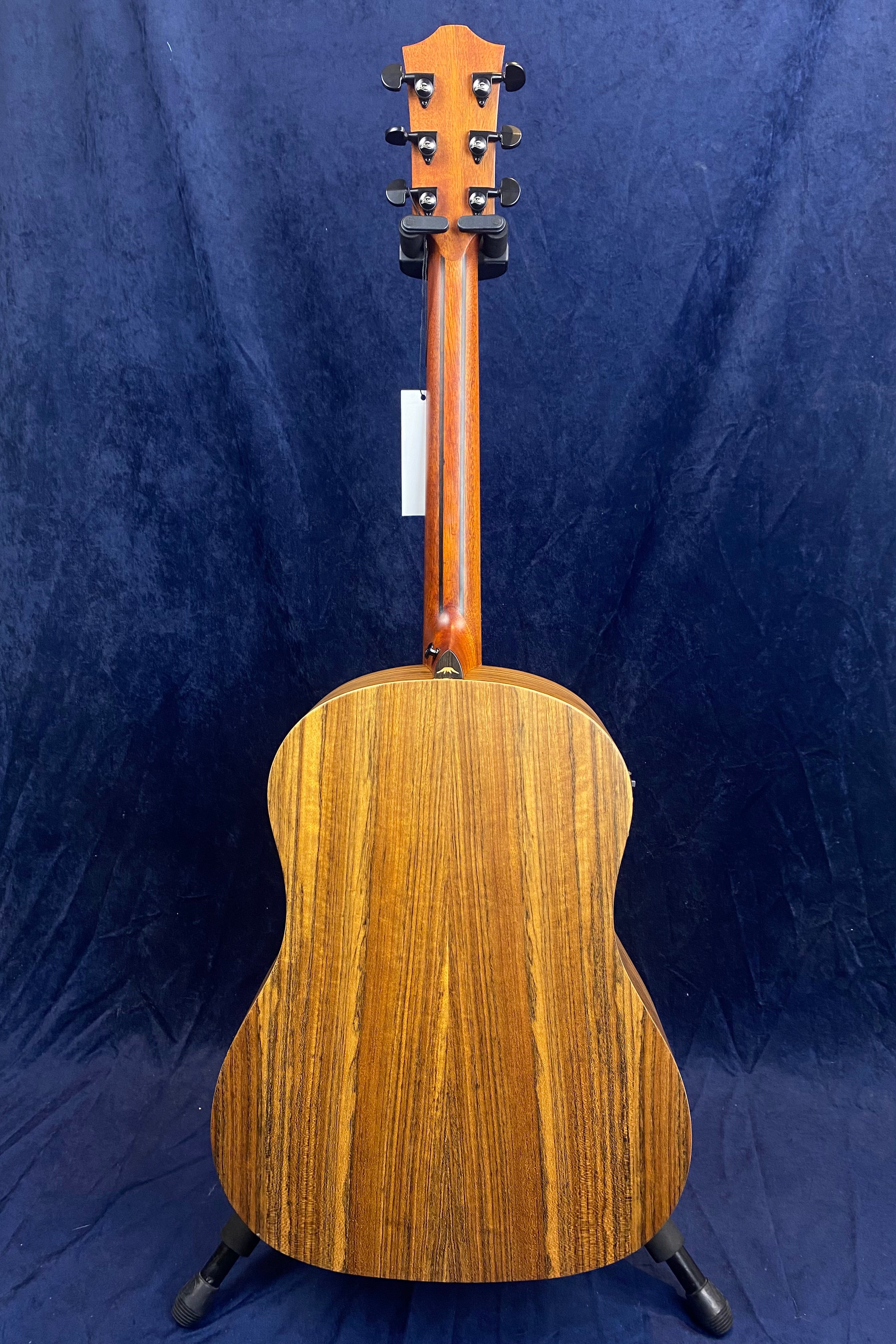 Acoustic guitar seen from the back, showing a smooth wooden finish with dark brown grain patterns and six tuning pegs on the headstock.